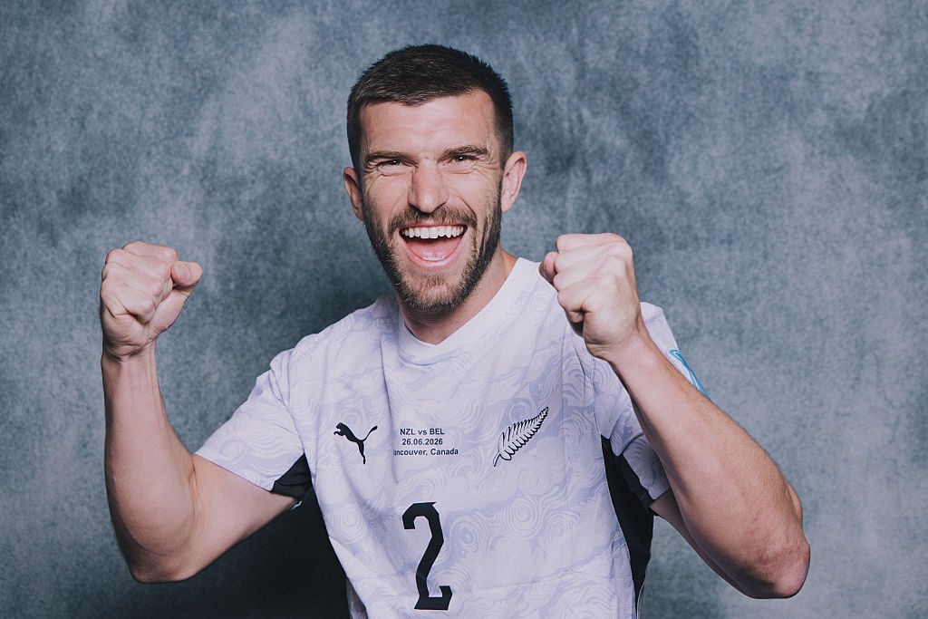 Homem branco barbudo, com cabelo curto e escuro, sorrindo amplamente e com os punhos cerrados, vestindo uma camisa de futebol branca com o número 2 e detalhes em preto, contra um fundo cinza texturizado