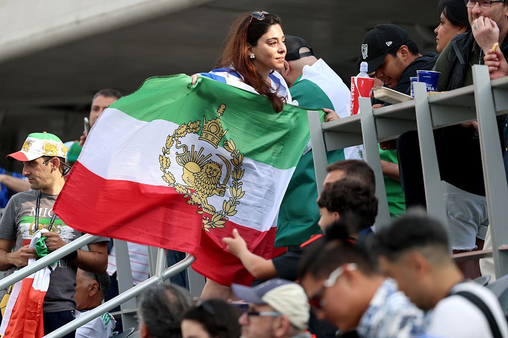Mulher de cabelo castanho e óculos de sol na cabeça segura uma bandeira do Irã com listras verde, branca e vermelha, e um leão dourado no centro, em meio a uma multidão em um estádio