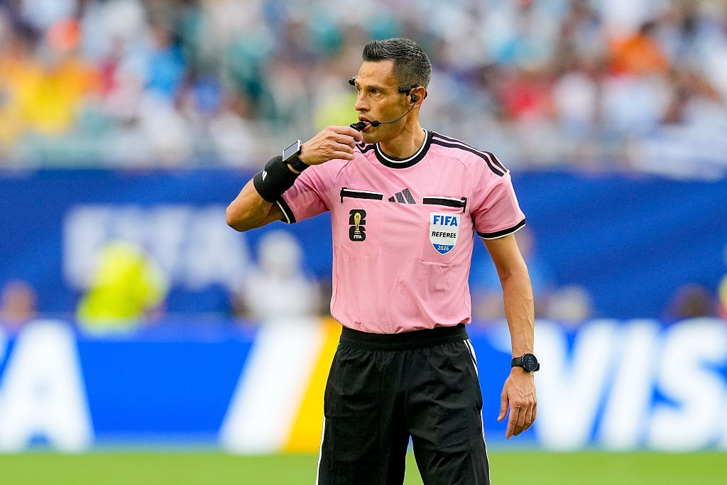 Árbitro de futebol masculino, com cabelo grisalho e uniforme rosa, apitando em campo. Ele usa um relógio inteligente no pulso esquerdo e um fone de ouvido.