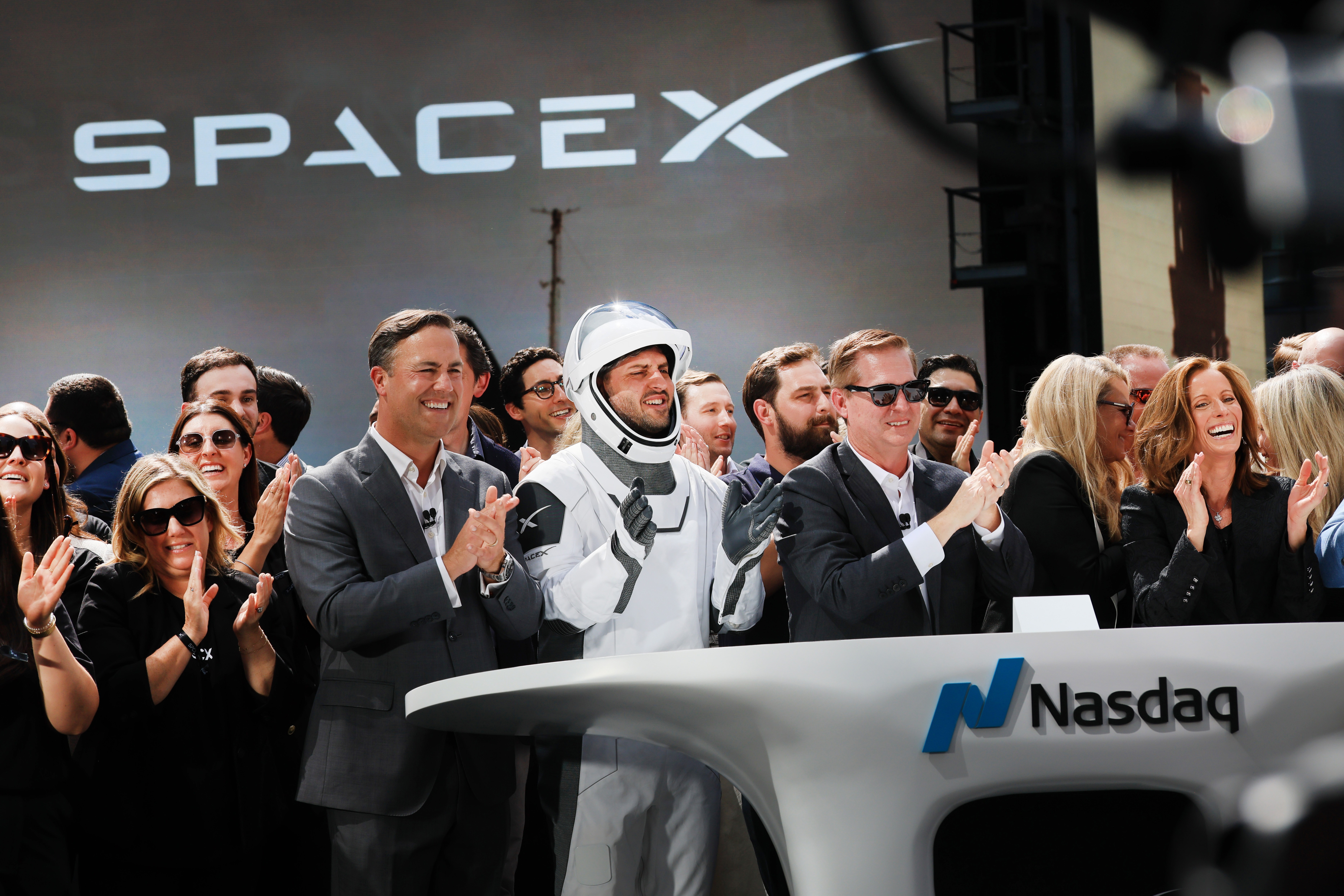 Pessoas aplaudem em evento da SpaceX e Nasdaq. Um homem sorri no centro, vestindo um traje espacial branco com capacete. À esquerda, um homem de terno cinza sorri e aplaude. À direita, outro homem de terno escuro e óculos de sol também aplaude. Mulheres e outros homens sorriem e batem palmas ao redor. Ao fundo, o logo SPACEX em tela grande. Na frente, um púlpito branco com o logo Nasdaq em azul e preto