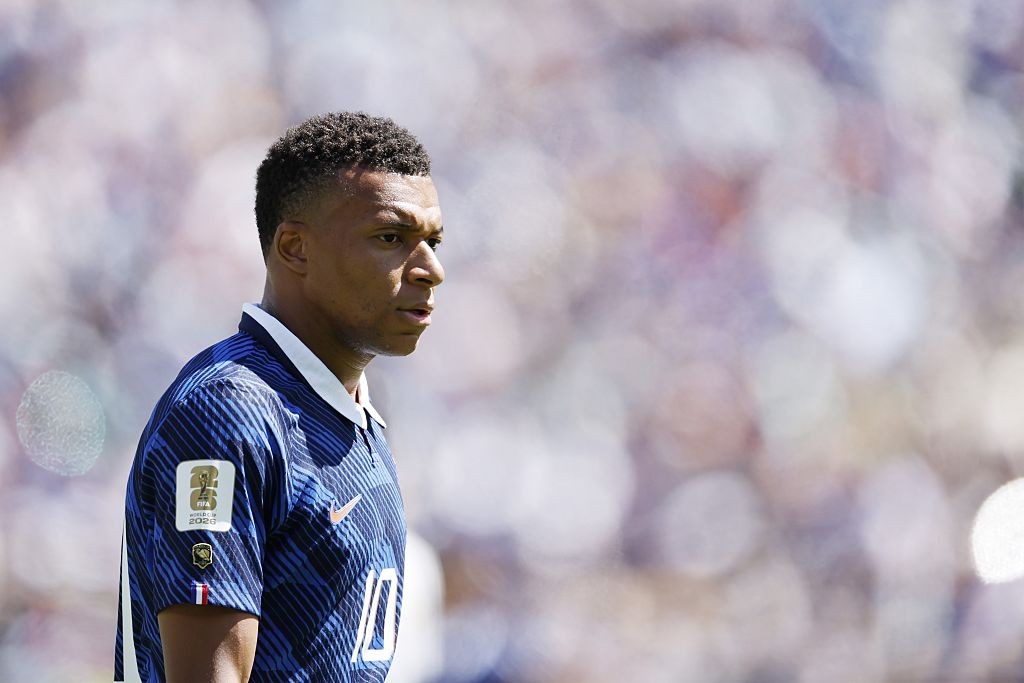 Kylian Mbappé, jogador de futebol, em perfil, usando camisa azul listrada com gola branca e o número 10, olhando para a direita, com fundo desfocado