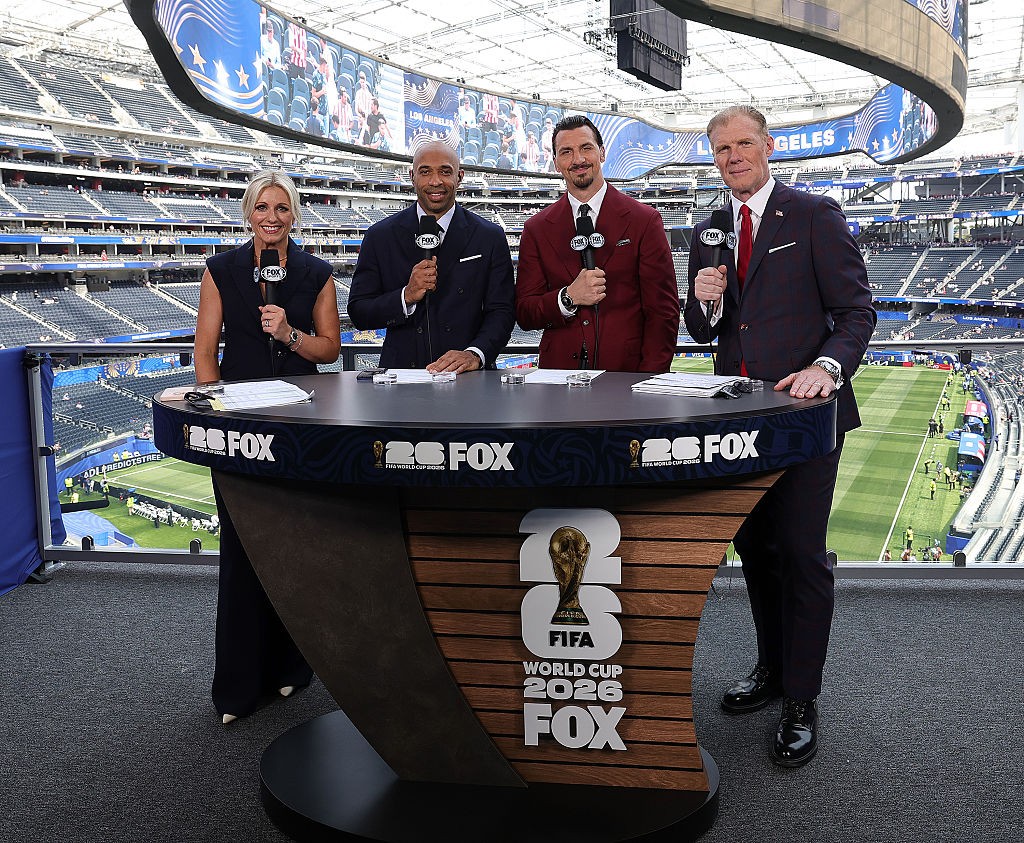 Quatro comentaristas esportivos, uma mulher e três homens, sorriem em uma bancada de transmissão da Fox, com o campo de futebol e as arquibancadas de um estádio ao fundo. A bancada exibe o logo da Copa do Mundo FIFA 2026
