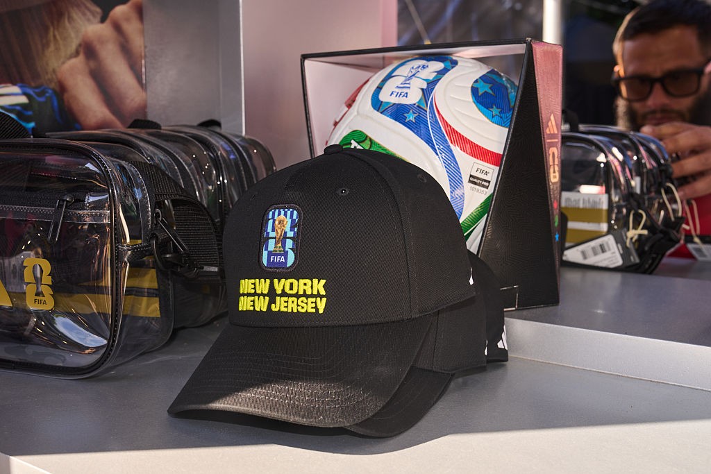 Bonés pretos com o logo da FIFA e New York New Jersey em amarelo, uma bola de futebol em caixa e bolsas transparentes com detalhes amarelos em uma loja. Um homem barbudo de óculos observa ao fundo