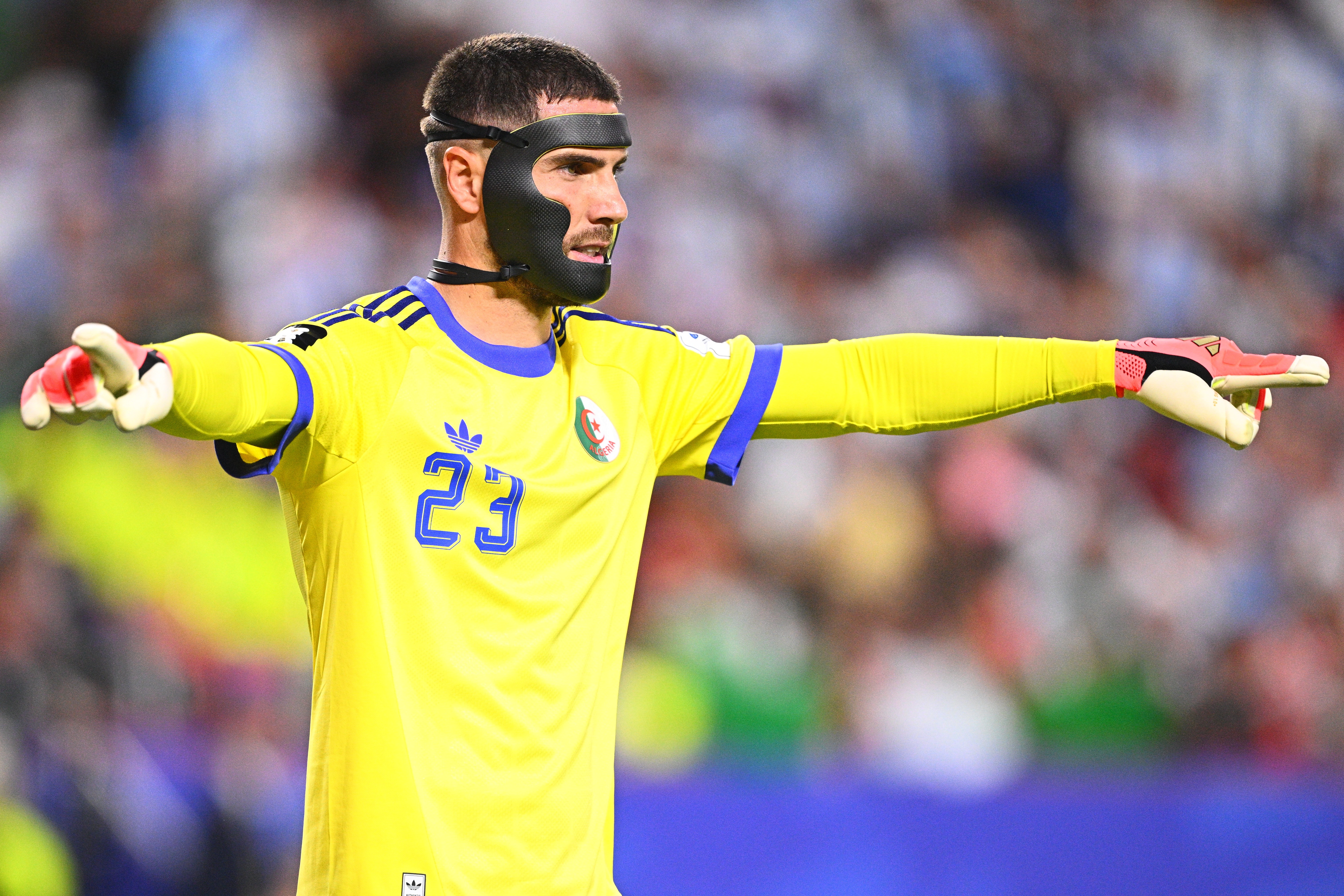 Goleiro com barba, vestindo camisa amarela com número 23 e luvas de goleiro, usando uma máscara protetora preta no rosto, apontando para a direita com o braço estendido em um campo de futebol