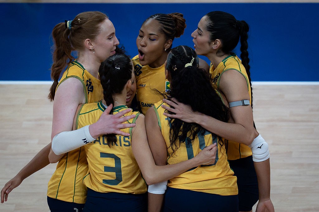 Cinco jogadoras de vôlei brasileiras, vestindo uniformes amarelos e azuis, abraçadas em quadra, com uma delas gritando de emoção