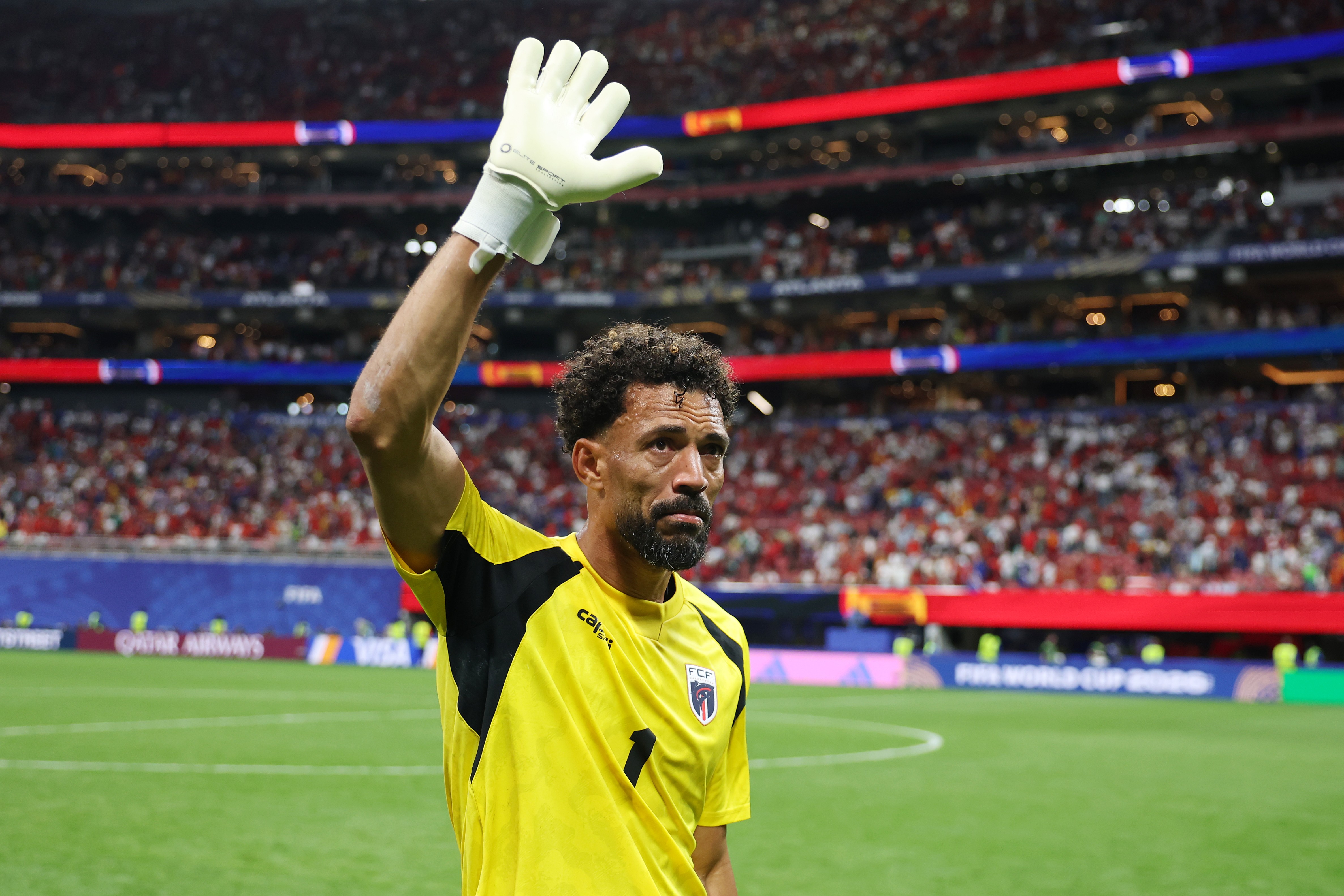 Goleiro de pele escura, barba e cabelo crespo, vestindo camisa amarela e luvas brancas, acena com a mão direita para a torcida em um estádio de futebol lotado