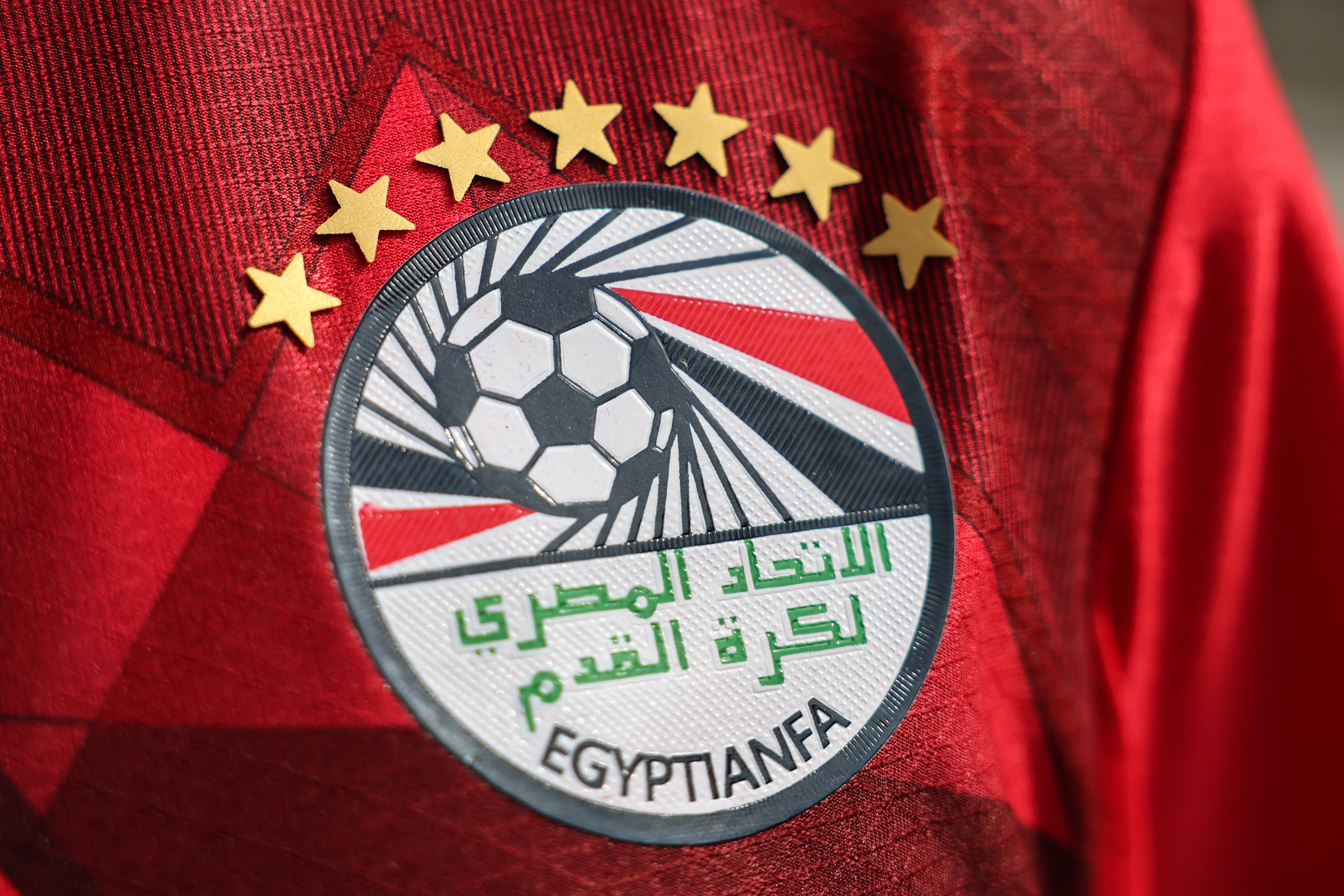 Emblema da Federação Egípcia de Futebol (EFA) em uma camisa vermelha, com seis estrelas douradas acima. O distintivo redondo tem uma bola de futebol no centro, com listras pretas e vermelhas em fundo branco, e texto em árabe e EGYPTIANFA abaixo