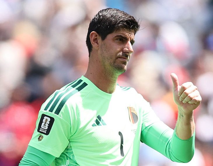 Goleiro de futebol masculino, pele clara e barba rala, vestindo uniforme verde-limão com detalhes em verde escuro, segurando luvas brancas na mão esquerda e fazendo sinal de positivo com a direita, em campo de futebol