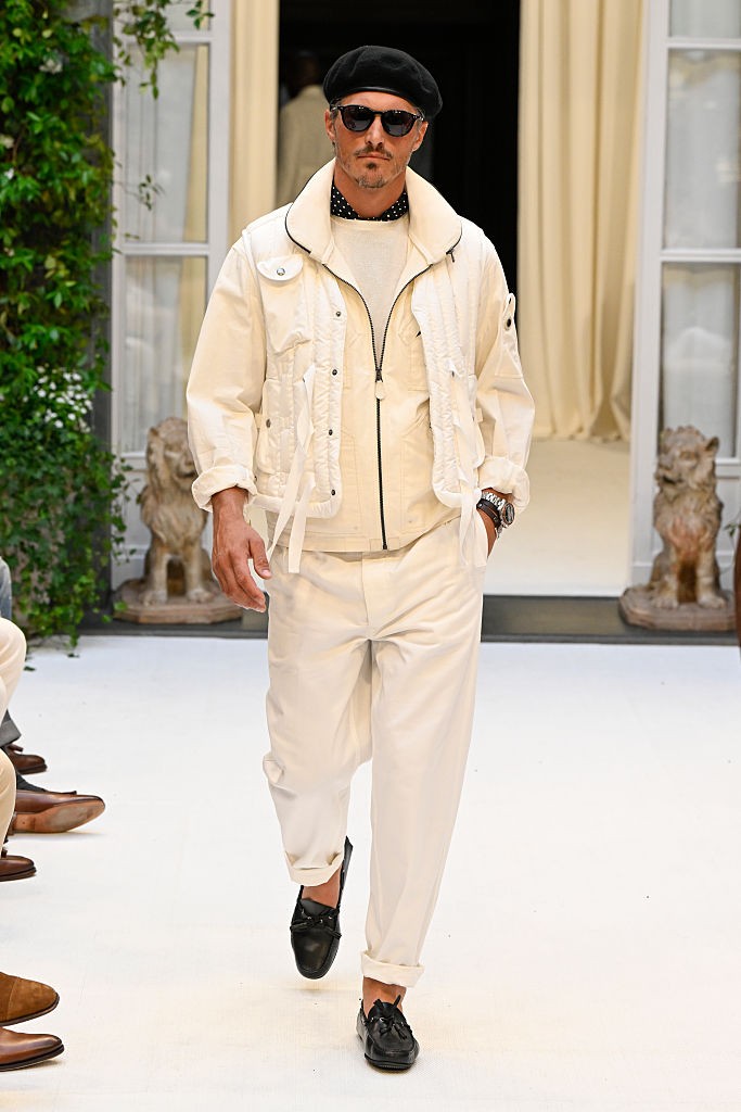 Homem branco de barba e bigode, usando boina preta, óculos escuros, jaqueta e calça creme, suéter branco e sapatos pretos, desfilando em passarela branca com estátuas de leões e plantas verdes ao fundo