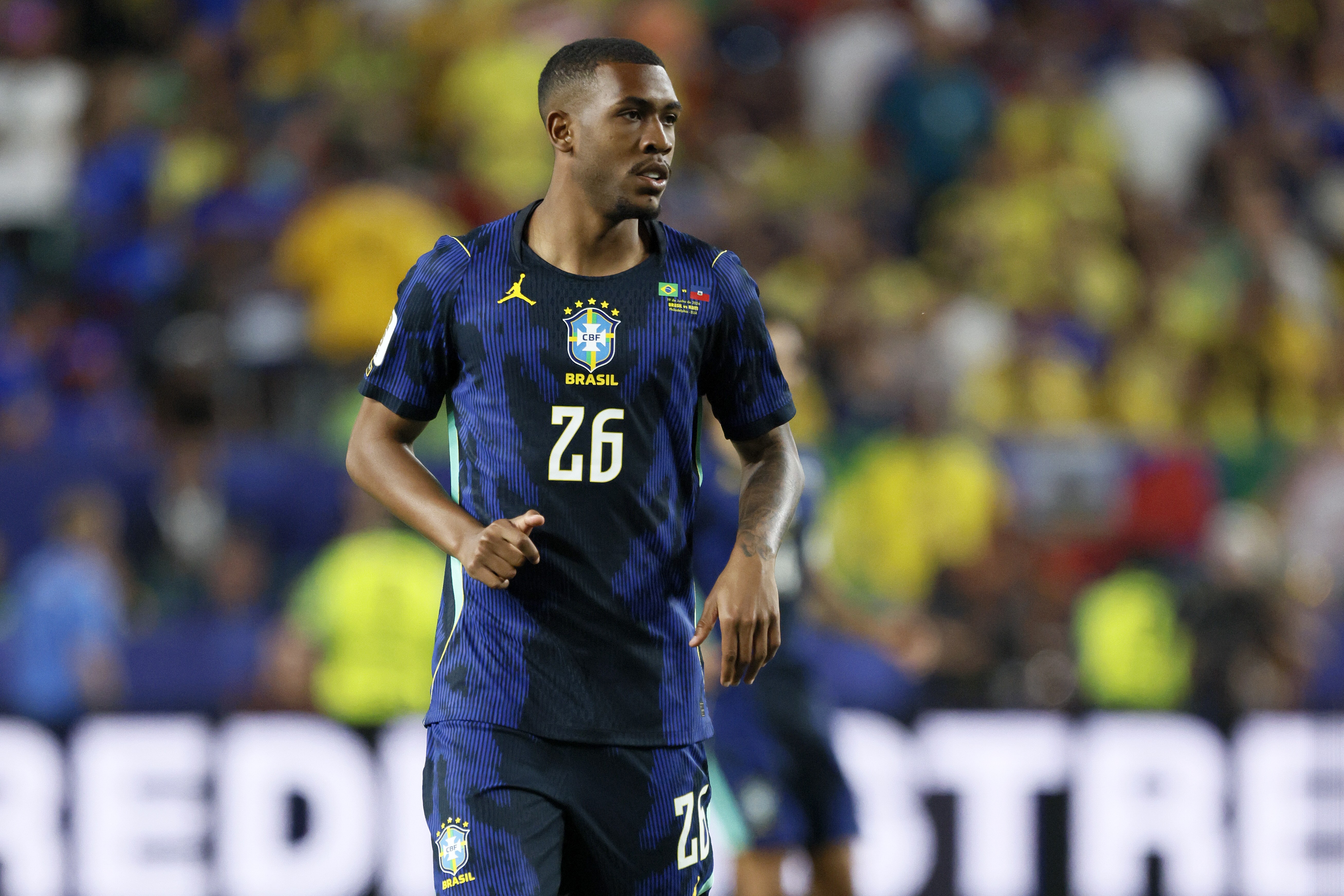 Jogador de futebol brasileiro, pele escura, cabelo curto, vestindo camisa azul escura com listras verticais e o número 26 em branco, em campo com torcida desfocada ao fundo