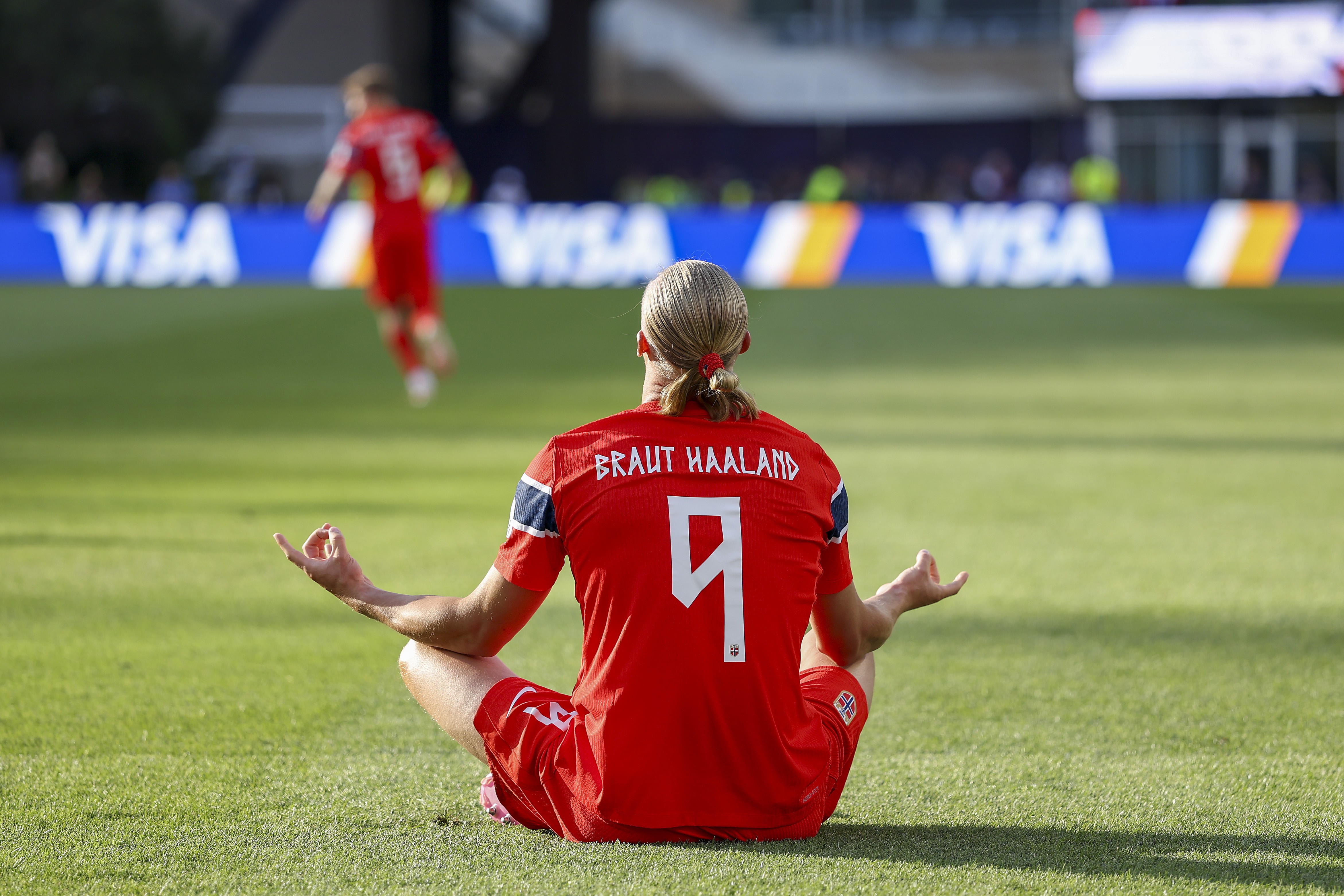 Jogador de futebol de costas, com camisa vermelha número 9 e nome BRAUT HAALAND, sentado em posição de lótus no gramado verde, meditando. Ao fundo, outro jogador desfocado e placas de publicidade VISA
