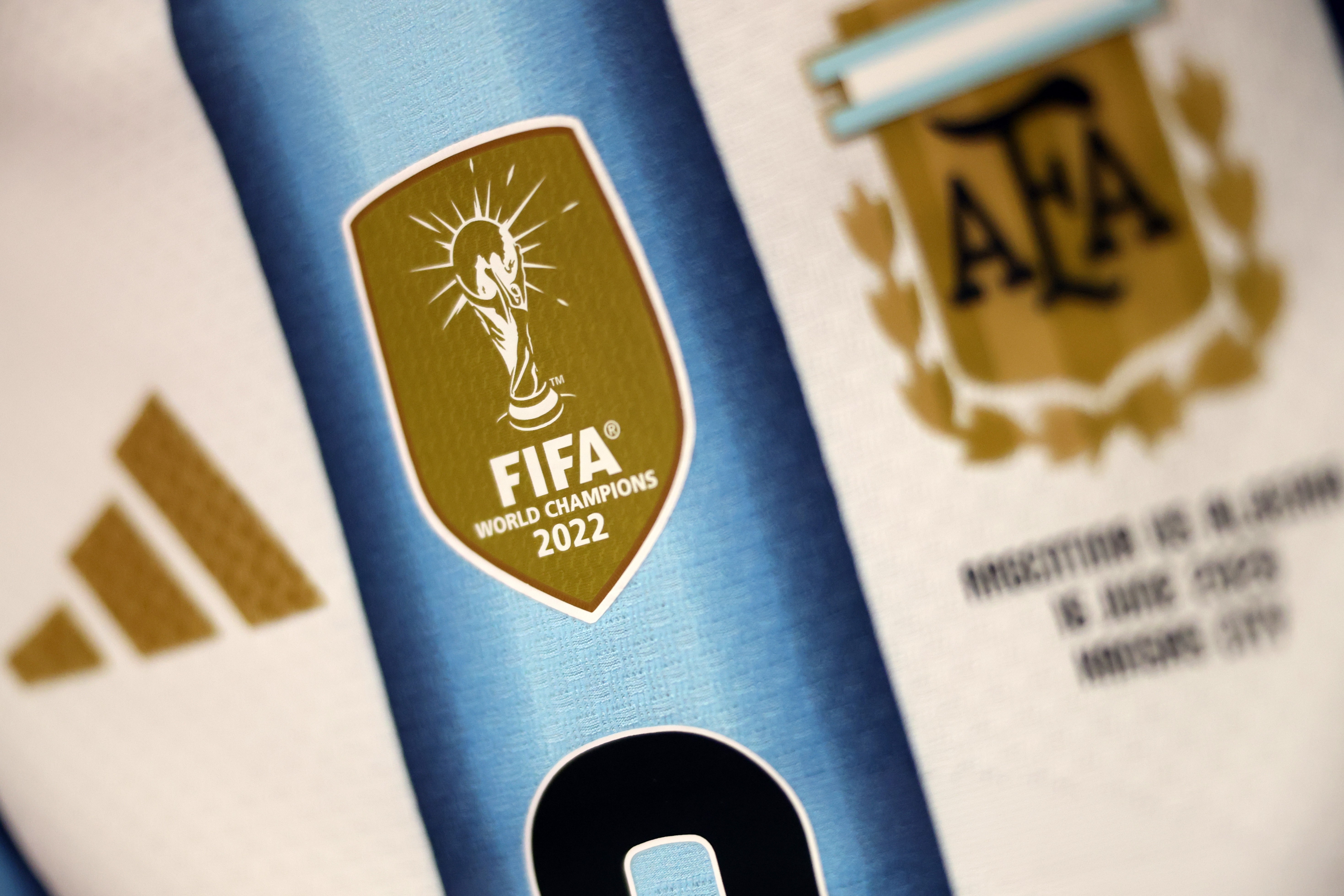 Detalhe da camisa da seleção argentina, mostrando o escudo dourado da FIFA World Champions 2022 com a taça, o logo da Adidas e o escudo da AFA