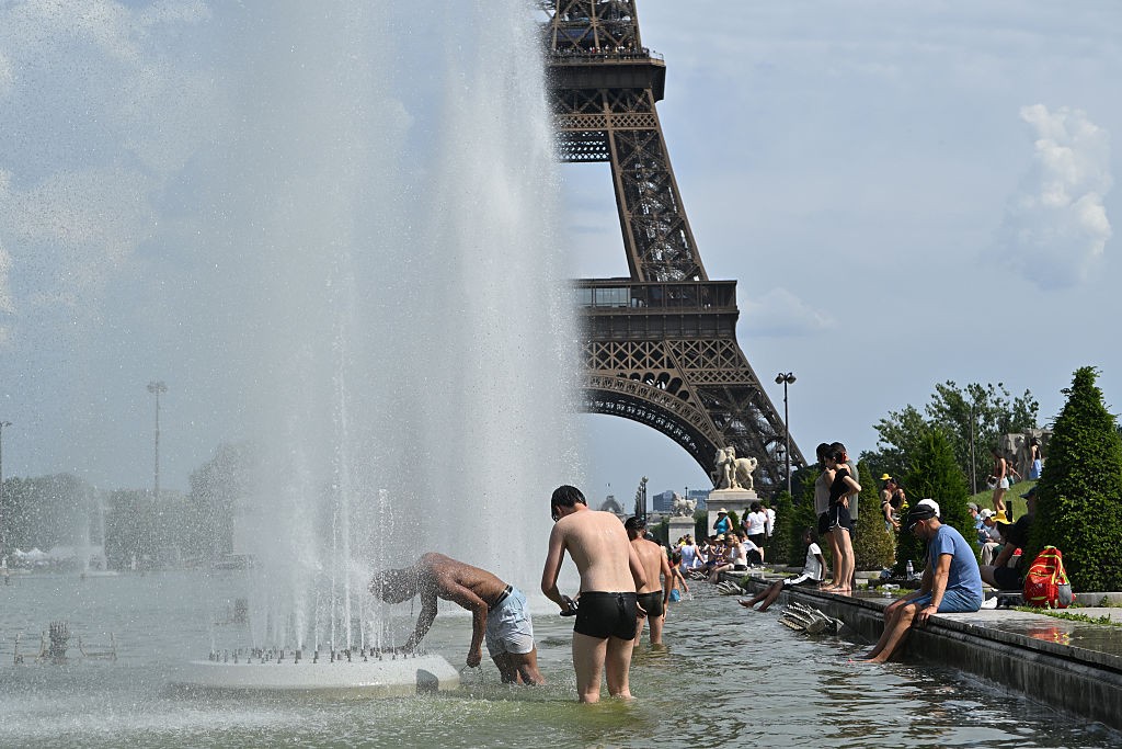 Pessoas se refrescam em uma fonte com jatos dágua em primeiro plano, enquanto a Torre Eiffel se ergue ao fundo, sob um céu parcialmente nublado. Alguns homens estão dentro da água, um deles se curvando para molhar a cabeça. Outras pessoas sentam-se na borda da fonte, com os pés na água, ou caminham ao redor. O ambiente é de lazer e alívio do calor.