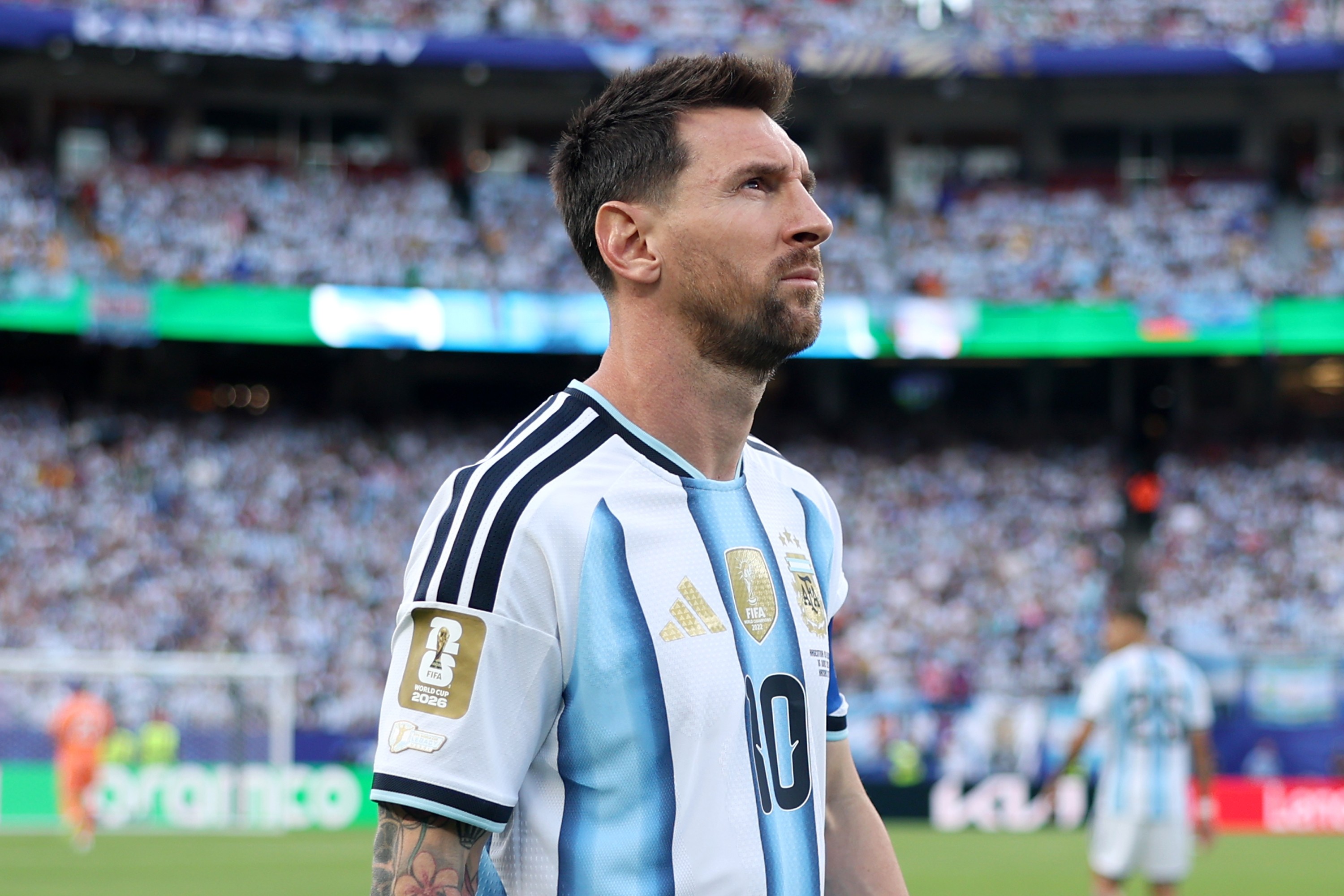 Lionel Messi, com barba e cabelo curto, veste a camisa branca e azul-celeste da Argentina, número 10, olhando para cima e para a direita em um estádio lotado