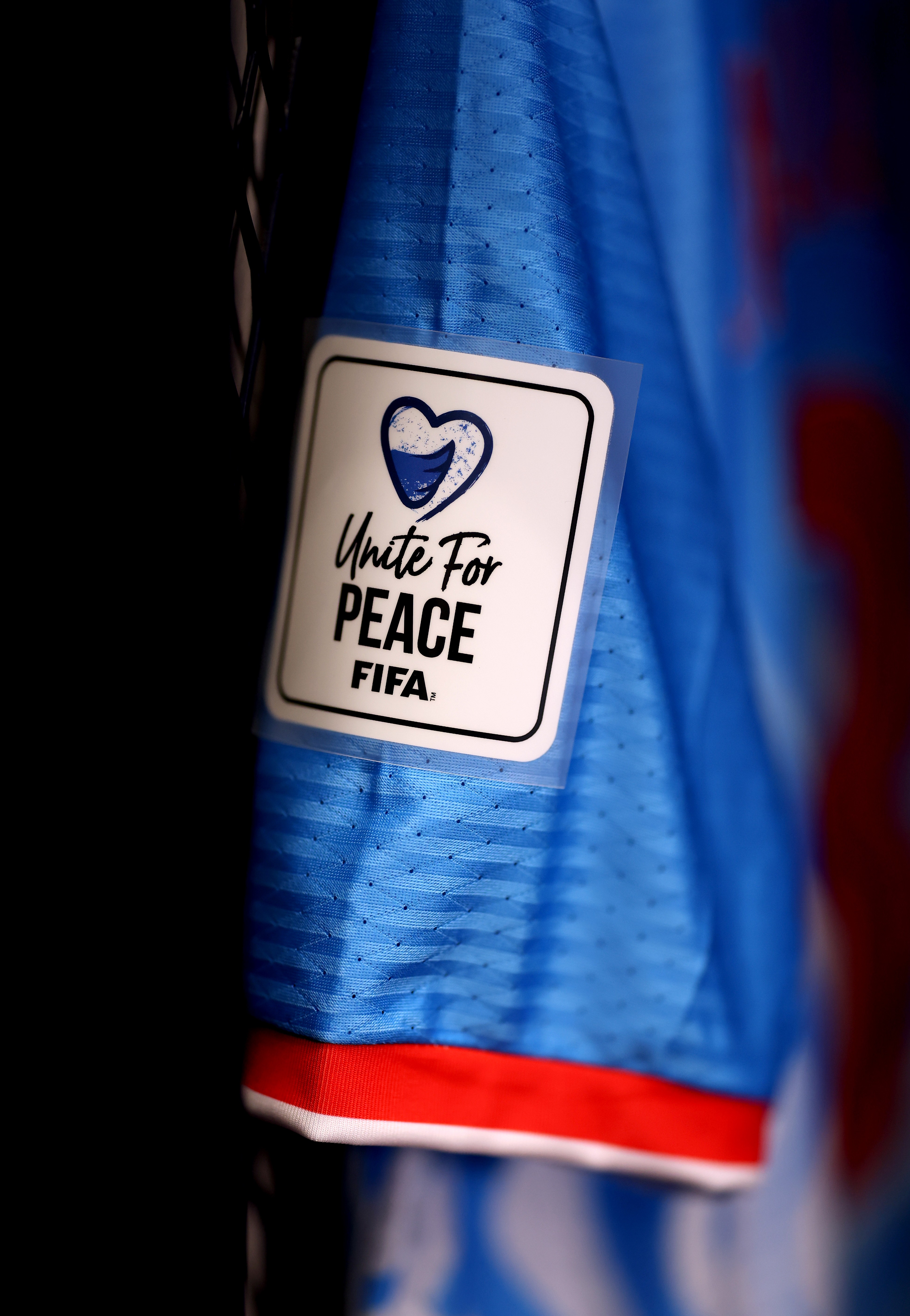 Detalhe de uma camisa de futebol azul com um patch branco retangular na manga, exibindo um coração azul e a frase Unite For PEACE FIFA em preto