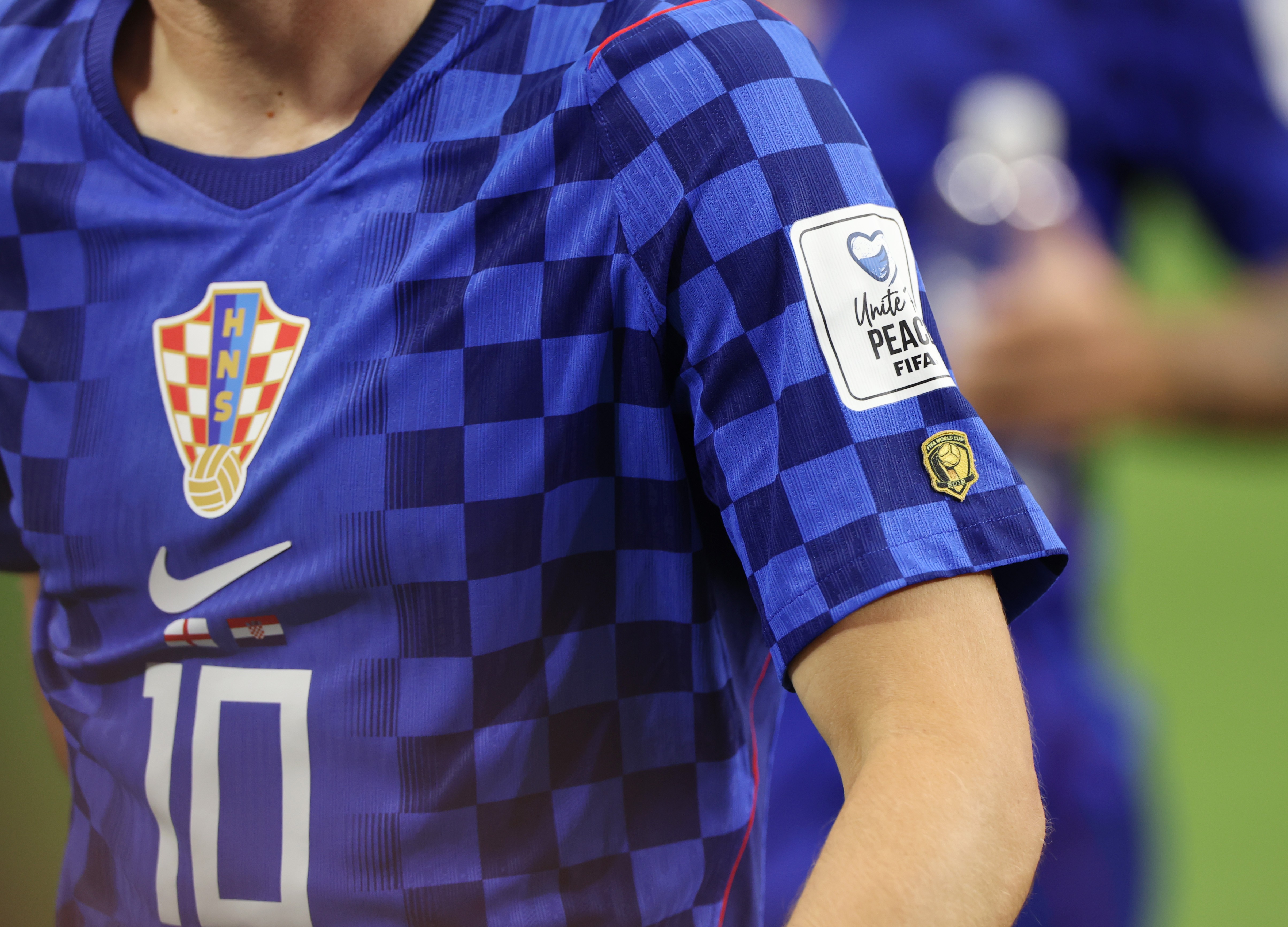 Camisa de futebol azul e xadrez com detalhes em azul escuro, escudo da Croácia, logo da Nike e número 10. Na manga direita, um patch branco Unite! Peace FIFA e um emblema dourado