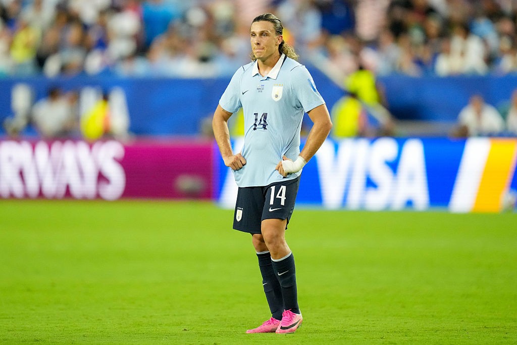 Jogador de futebol com camisa azul-clara e shorts pretos, número 14, em pé no gramado verde de um estádio, mãos na cintura, olhando para frente. Ele tem cabelos longos e loiros presos para trás, e uma faixa branca no punho esquerdo. Ao fundo, placas de publicidade com AIRWAYS e VISA e torcedores borrados.