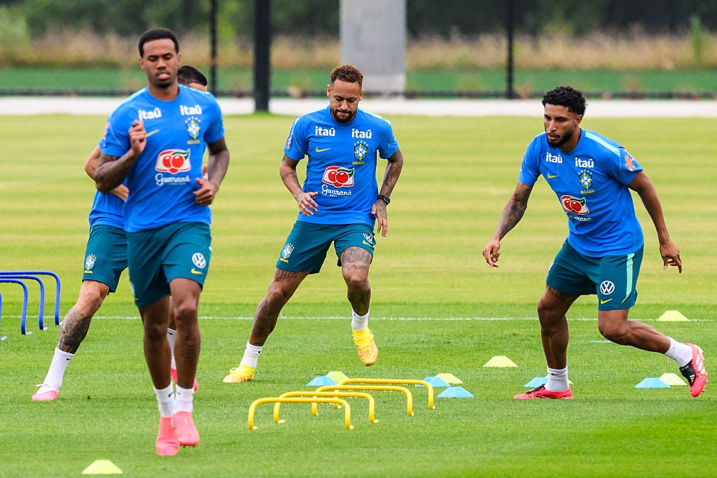 Três jogadores de futebol, Neymar Jr. ao centro, correm em campo de grama verde, usando uniformes azuis e shorts verde-escuros. Eles participam de um treino com pequenos cones e barreiras amarelas no chão