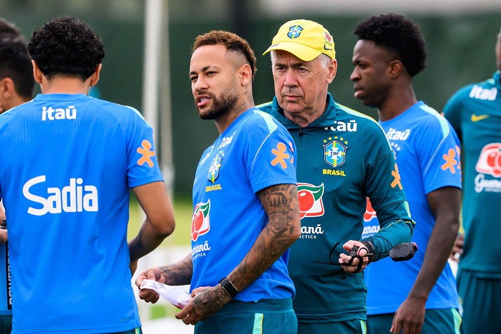 Neymar, de barba e cabelo curto, veste camisa azul da seleção brasileira, olhando para a esquerda. Ao lado, um homem mais velho de boné amarelo e camisa verde da seleção, segura um objeto preto. Outros jogadores de camisa azul estão ao redor, em um campo de treino