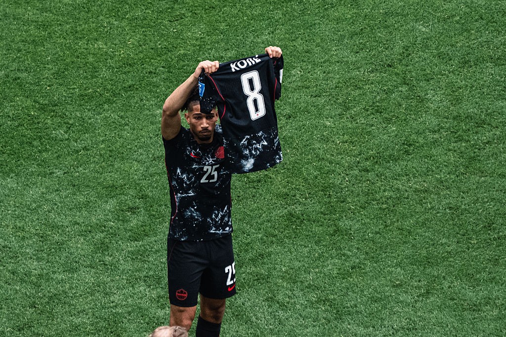 Jogador de futebol com barba, vestindo uniforme preto com detalhes brancos e número 25, ergue uma camisa preta com o nome KOBE e o número 8 em branco, em um campo de grama verde