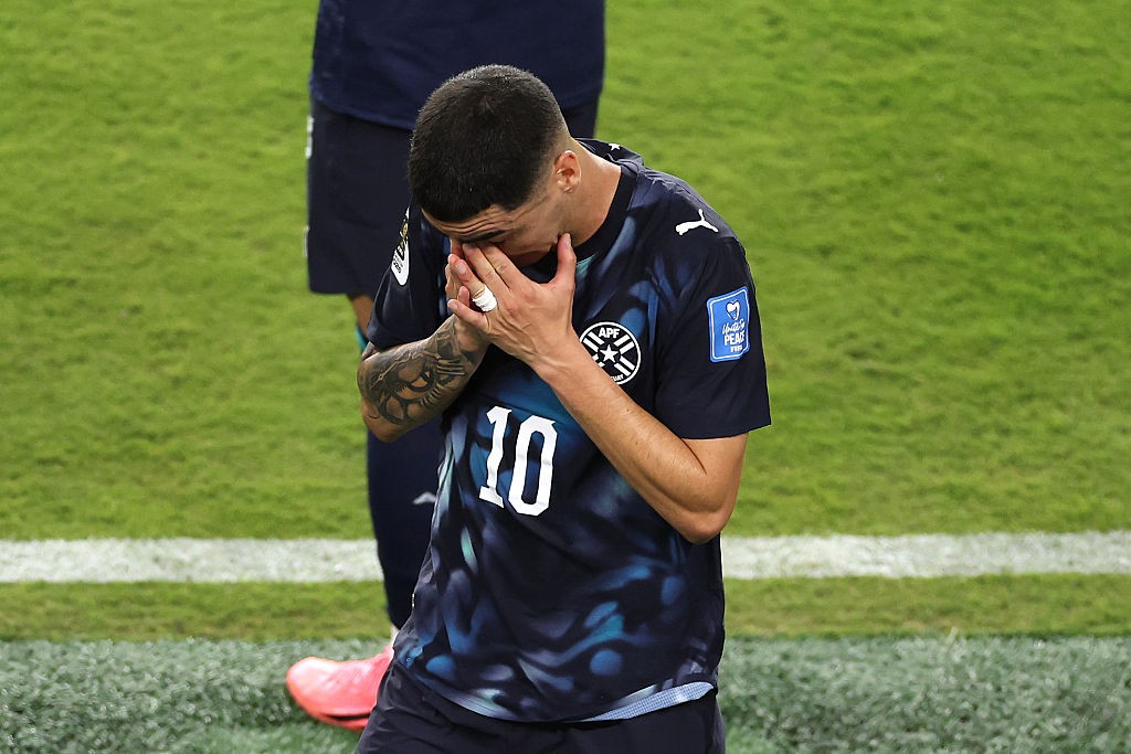 Jogador de futebol com camisa azul escura e número 10, cobrindo o rosto com as mãos, em campo verde. Ele usa uma faixa branca no dedo e tem tatuagens no braço esquerdo. Um jogador de uniforme escuro e chuteiras rosas aparece desfocado ao fundo