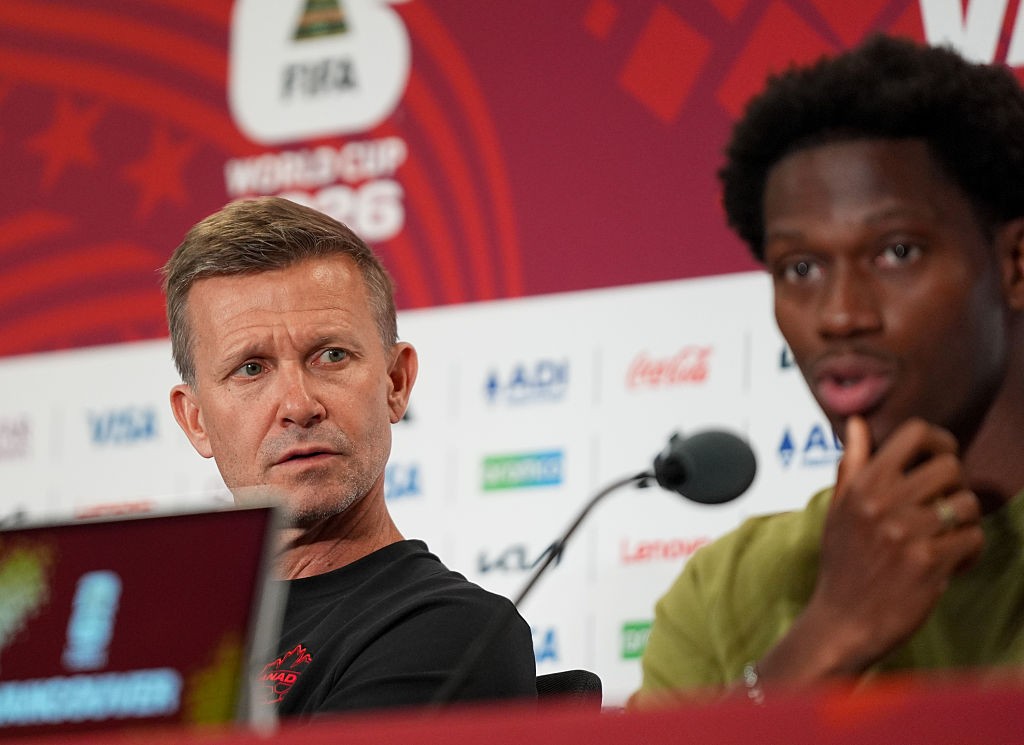 Um homem branco de cabelos curtos e grisalhos, vestindo camiseta preta, olha atentamente para frente em uma coletiva de imprensa. Ao fundo, um painel vermelho com o logo da FIFA World Cup 26. À direita, um homem negro com cabelo crespo e camiseta verde, desfocado, parece falar ou reagir a algo, com a mão perto do queixo
