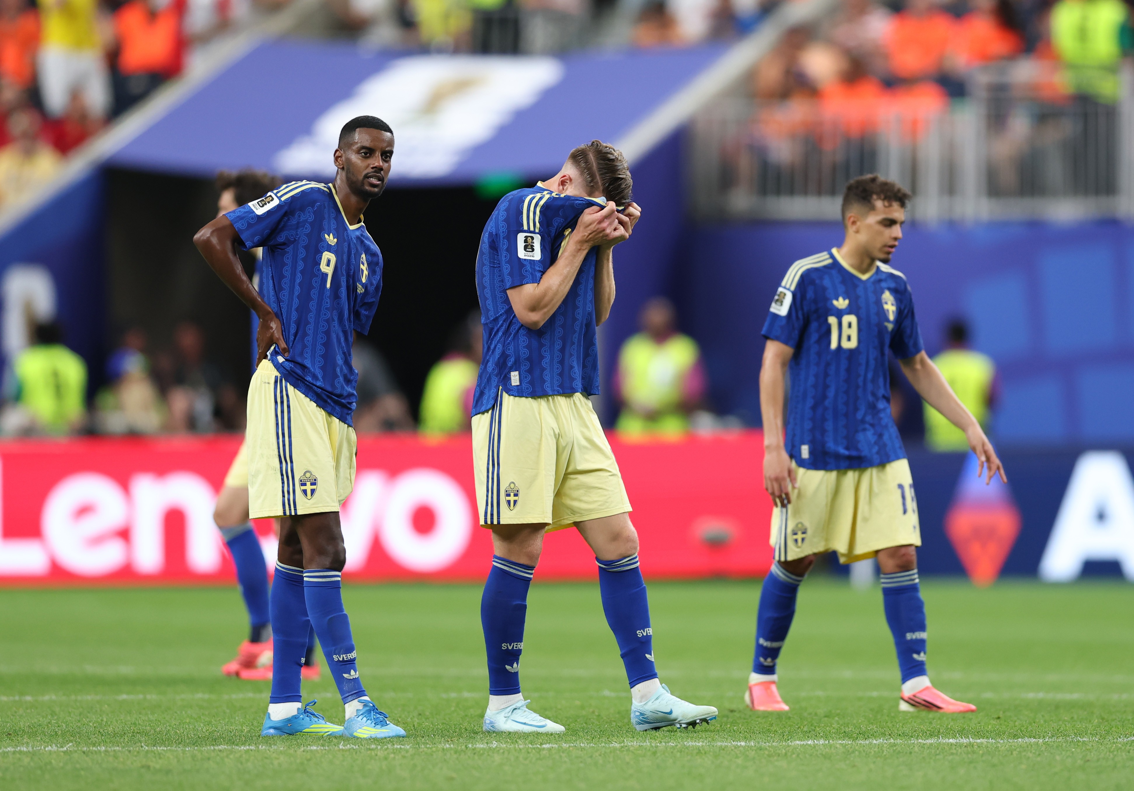 Três jogadores de futebol com uniformes azuis e shorts amarelos em campo. Um jogador à esquerda olha para frente, outro ao centro cobre o rosto com as mãos, e o terceiro à direita olha para baixo, com expressão de desânimo. Ao fundo, um banner vermelho com a palavra Lenovo em branco e torcedores borrados.