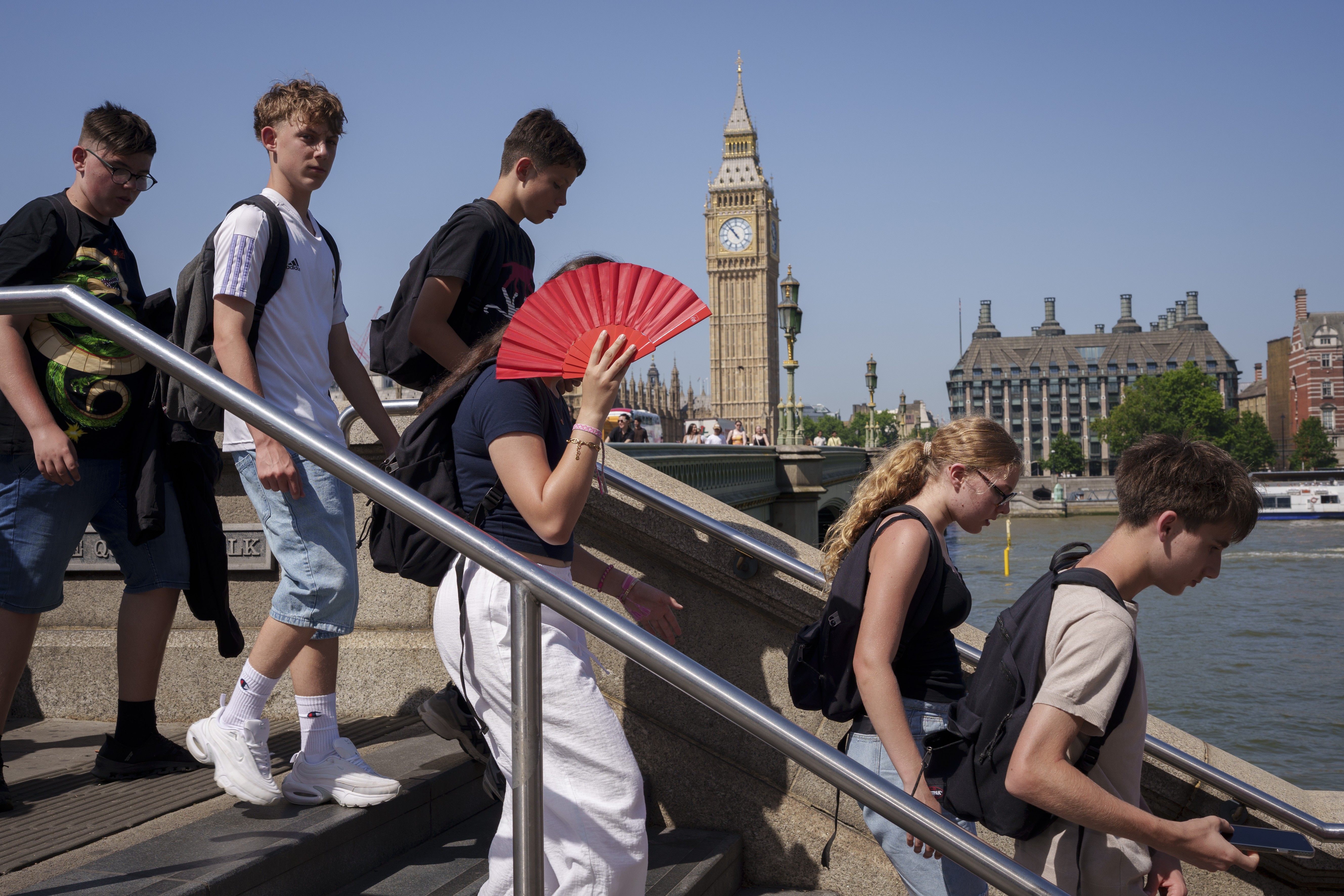 Grupo de jovens descendo escadas ao ar livre, com a torre do Big Ben e o rio Tâmisa ao fundo. Uma jovem no centro usa um leque vermelho para se refrescar. Todos vestem roupas casuais e mochilas, em um dia ensolarado