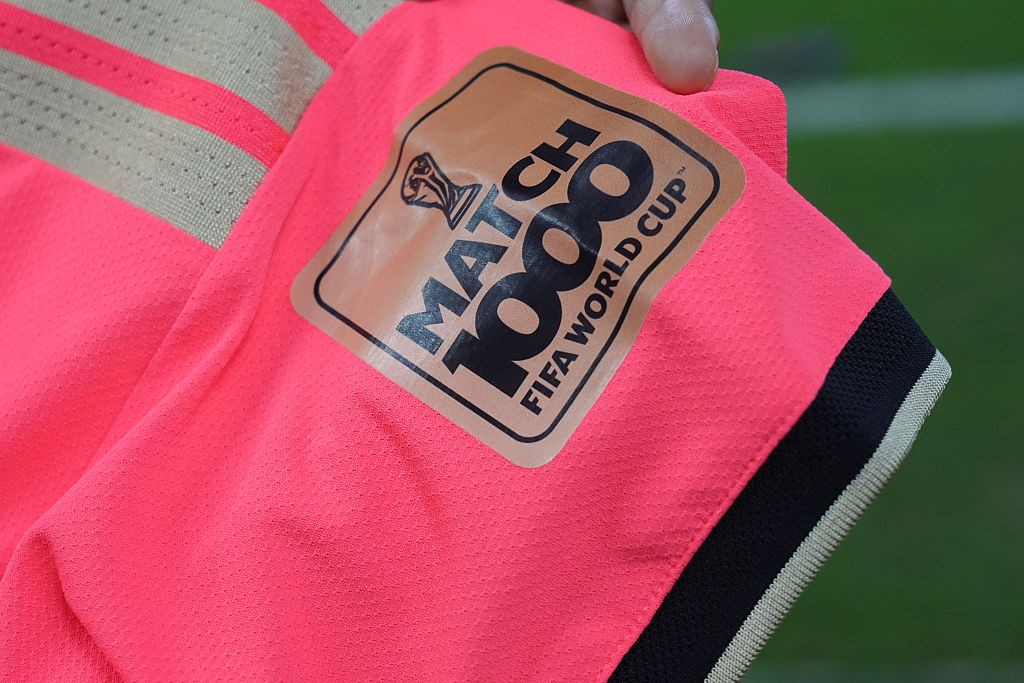 Detalhe de uma camisa de futebol rosa com listras cinzas e um patch marrom-claro na manga. O patch tem a inscrição MATCH 1000 FIFA WORLD CUP em preto, com um troféu da Copa do Mundo acima da palavra MATCH. Um dedo segura a camisa na parte superior. O fundo é um campo de grama verde desfocado