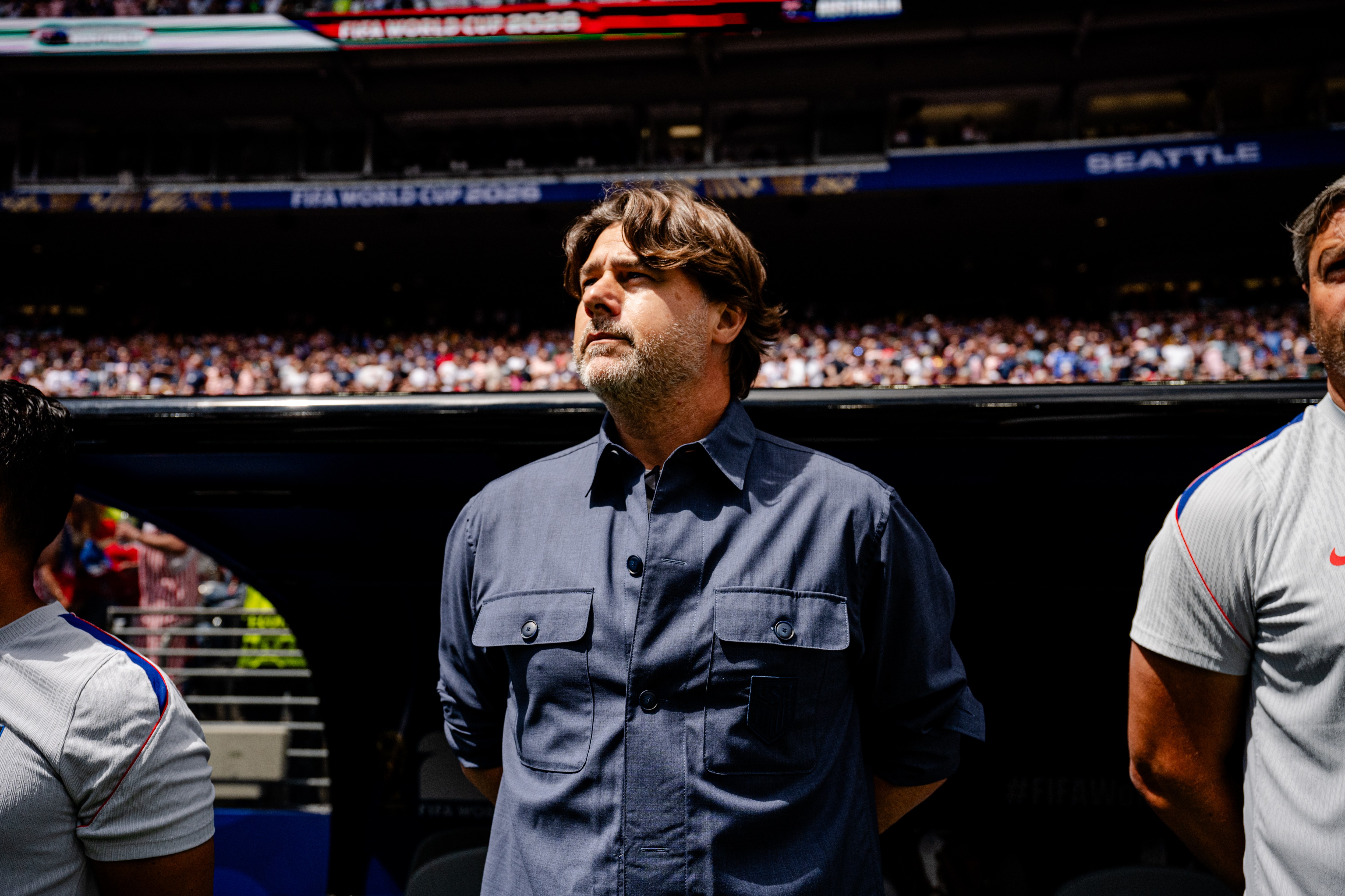 Homem de barba e cabelo castanho, vestindo camisa azul-marinho com bolsos frontais, olhando para cima em um estádio lotado. Ao fundo, um telão exibe FIFA WORLD CUP 2026 e SEATTLE.
