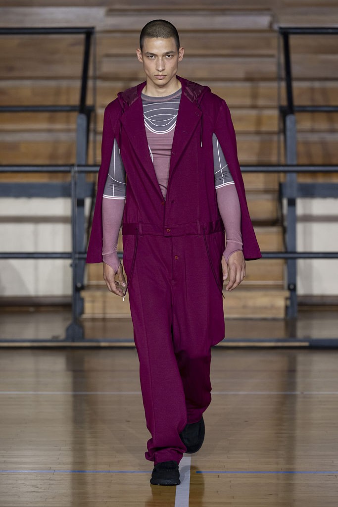 Homem de pele clara e cabelo raspado, vestindo um macacão magenta com capuz e uma blusa cinza com listras brancas, desfila em uma passarela de madeira com marcações esportivas
