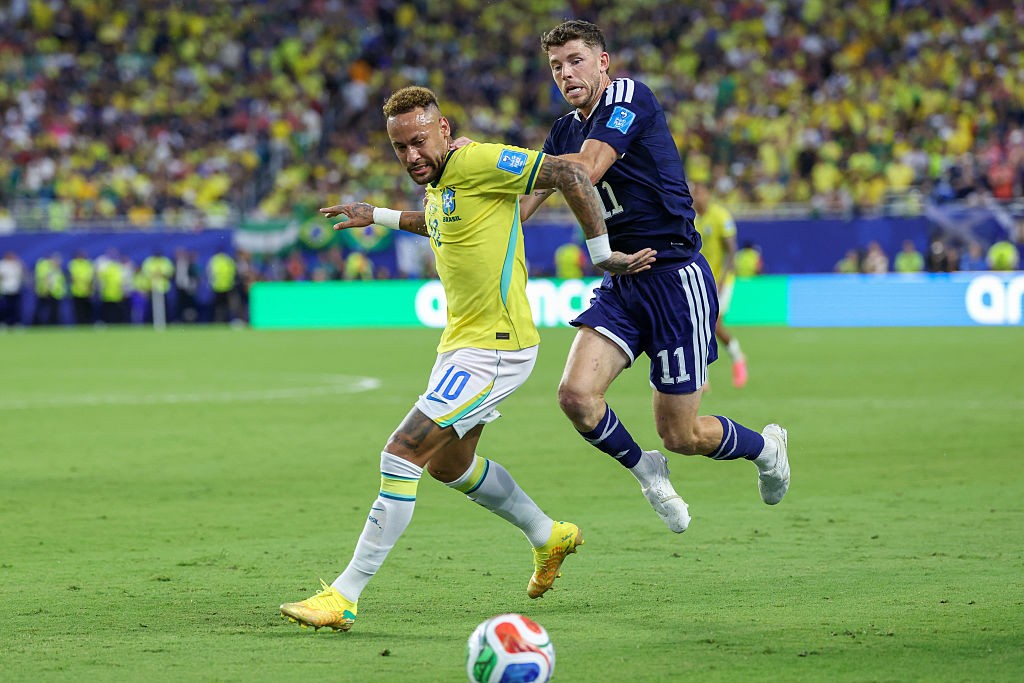 Neymar, com camisa amarela e shorts brancos, dribla um jogador de camisa azul marinho e shorts azuis, em campo de futebol com torcida ao fundo