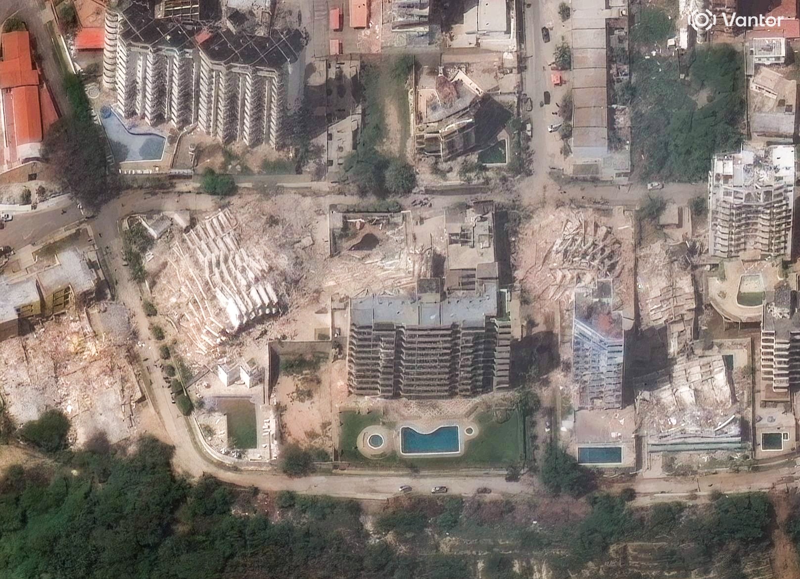 Vista aérea de uma área urbana com edifícios residenciais, alguns intactos com piscinas, e outros completamente destruídos, reduzidos a escombros. Há ruas visíveis e áreas arborizadas na parte inferior da imagem, indicando um cenário de devastação em meio a construções. O logo da Vantor aparece no canto superior direito.