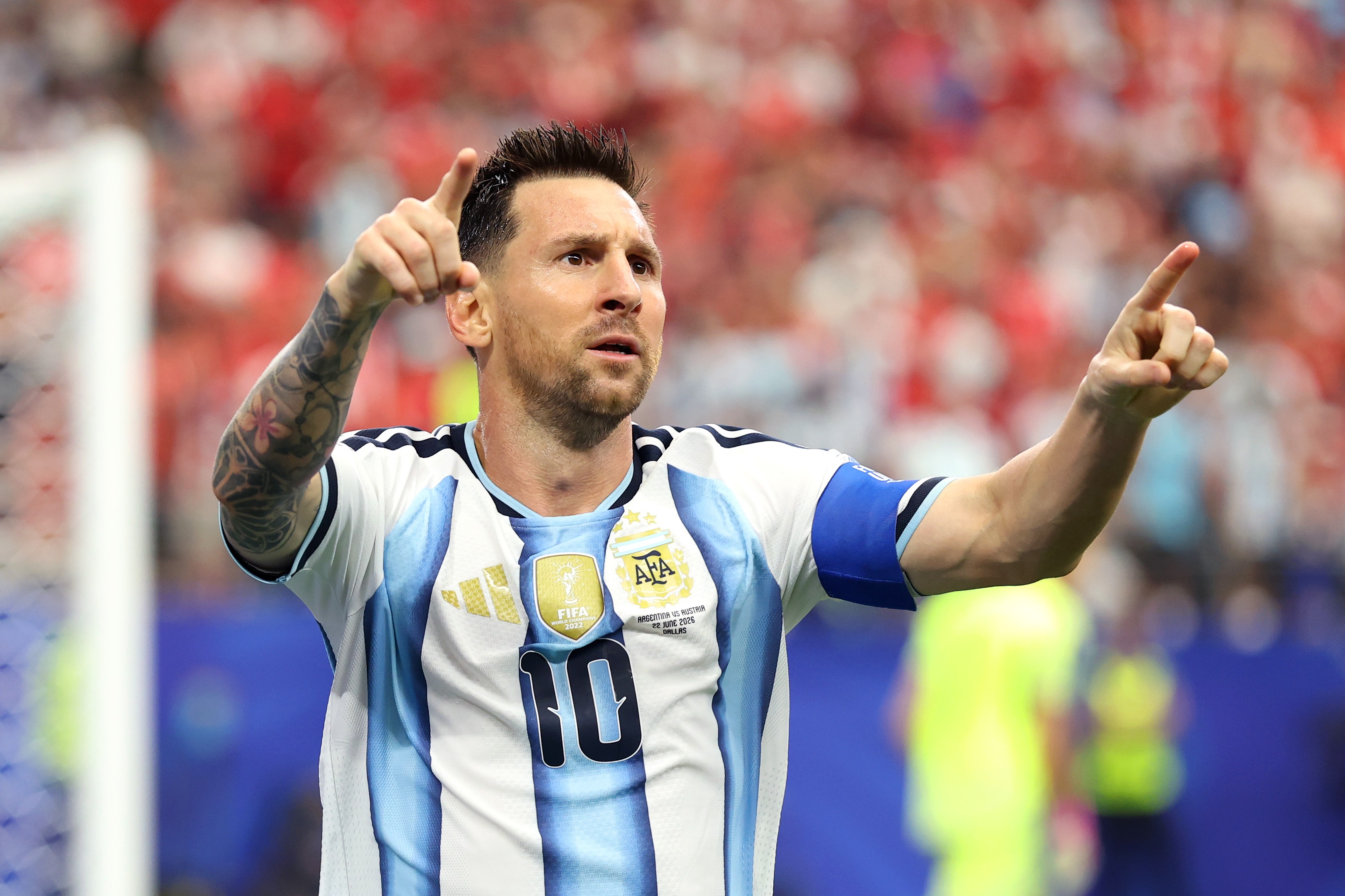Lionel Messi, jogador de futebol argentino, com barba e cabelo escuro, vestindo a camisa listrada azul e branca da Argentina com o número 10, aponta para cima com as duas mãos, em um estádio lotado