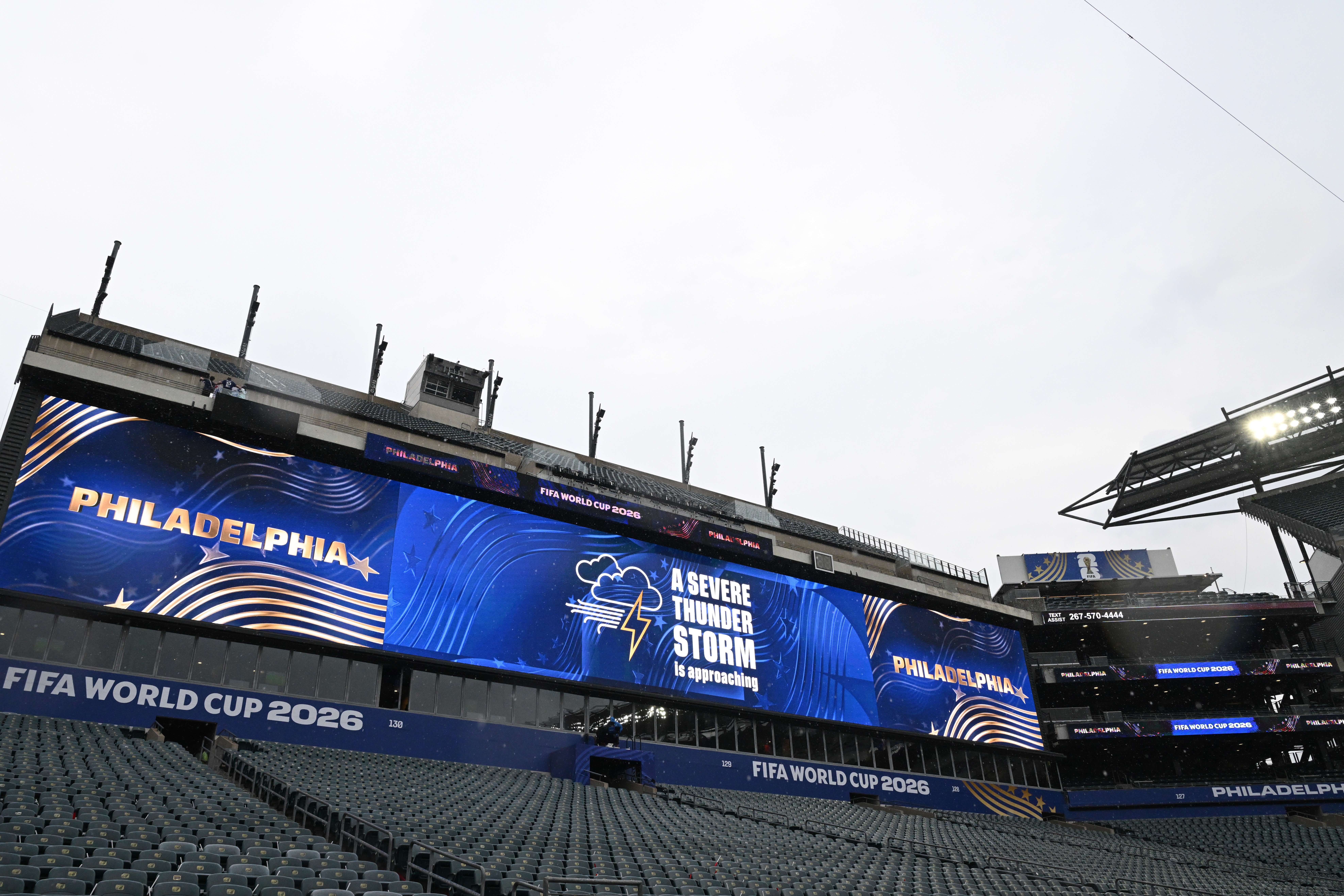 Telões de um estádio de futebol exibem Philadelphia e um alerta de tempestade severa com nuvem e raio, sob um céu claro. As arquibancadas escuras estão vazias, e uma faixa azul abaixo dos telões diz FIFA World Cup 2026