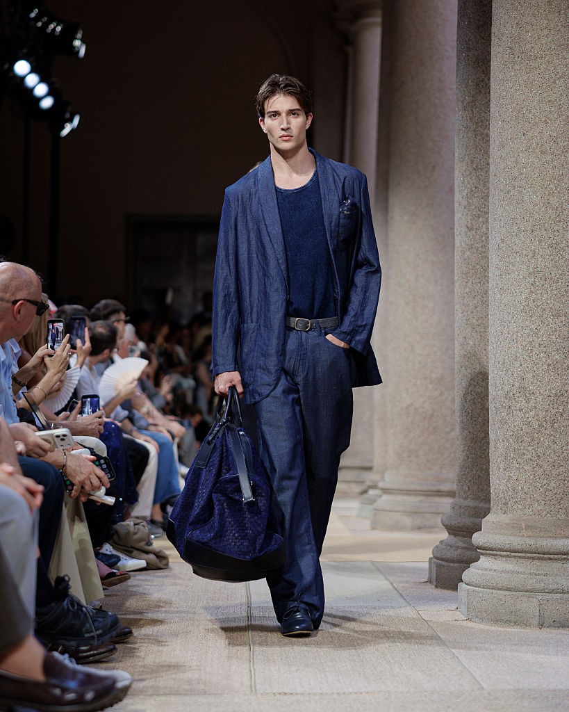 Modelo masculino jovem, com cabelo castanho e expressão séria, desfila em passarela. Veste blazer e calça jeans azul-marinho, camiseta azul escura e cinto preto, segurando uma bolsa de viagem azul escura. Ao fundo, colunas de pedra e público assistindo, alguns filmando com celulares