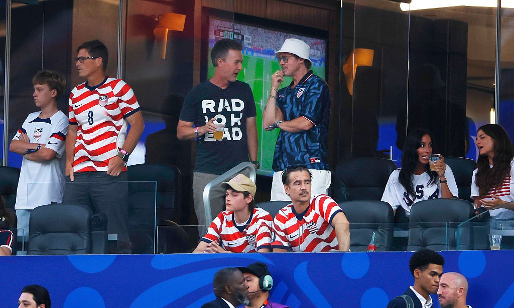 Pessoas assistindo a um jogo de futebol em um camarote. À esquerda, um homem de óculos e camisa listrada vermelha e branca com o número 8, ao lado de um garoto de camisa branca. No centro, dois homens conversam, um de camiseta preta e outro de chapéu bucket branco e camisa azul escura. À direita, duas mulheres sentadas, uma de camisa branca com USA e outra de camisa listrada. Abaixo, dois homens sentados com camisas listradas e, na parte inferior, outras pessoas em um ambiente azul