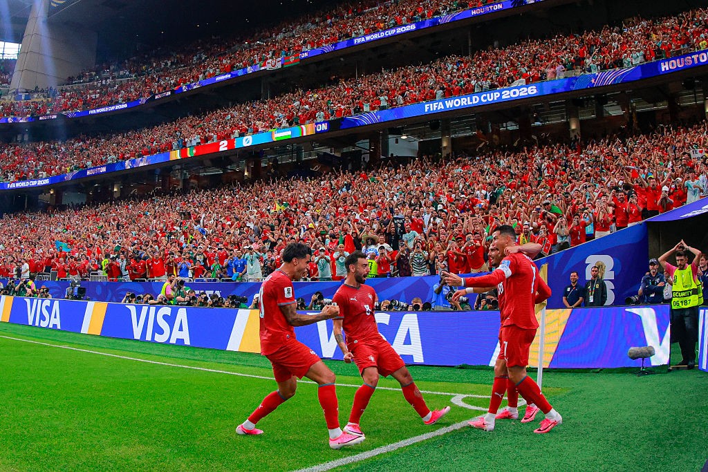 Quatro jogadores de futebol em uniformes vermelhos celebram um gol no campo verde, com um estádio lotado de torcedores em vermelho ao fundo. Um jogador abraça outro, enquanto os outros dois se aproximam, sorrindo. Placas de publicidade VISA e FIFA WORLD CUP 2026 são visíveis.