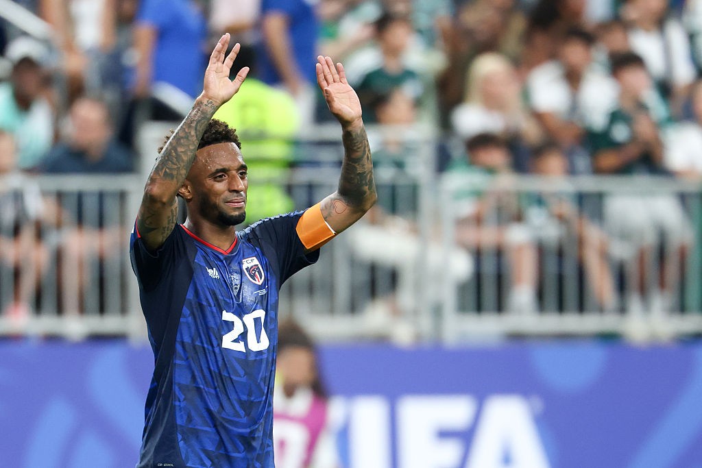 Jogador de futebol negro, com barba e cabelo curto, vestindo camisa azul com o número 20, braços tatuados levantados e mãos abertas, acenando para a torcida em um estádio, com a arquibancada desfocada ao fundo