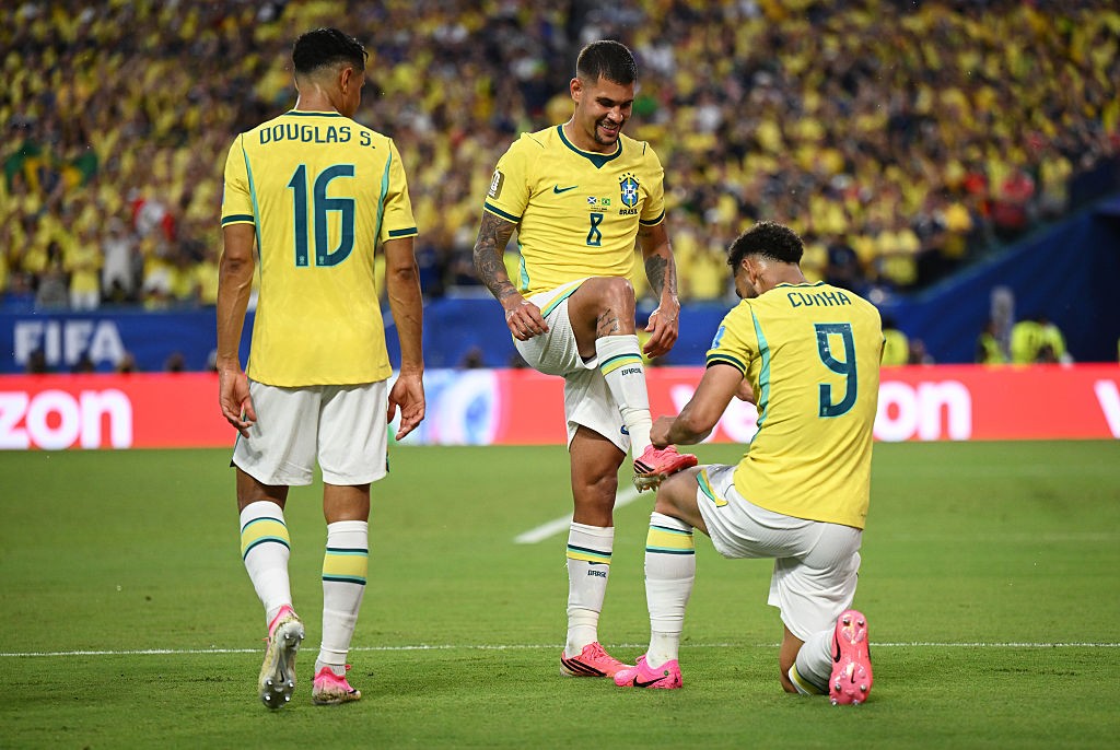 Três jogadores de futebol brasileiros em campo, vestindo uniformes amarelos e brancos. Um jogador ajoelhado amarra a chuteira rosa de outro, que sorri. O terceiro jogador, de costas, observa a cena. A arquibancada lotada de torcedores amarelos e um letreiro vermelho com FIFA e zon ao fundo completam o cenário