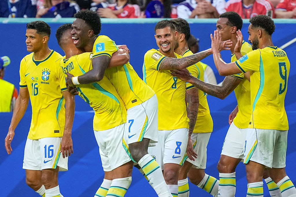 Jogadores de futebol brasileiros em uniformes amarelos e brancos celebram um gol, abraçando-se e sorrindo em campo azul