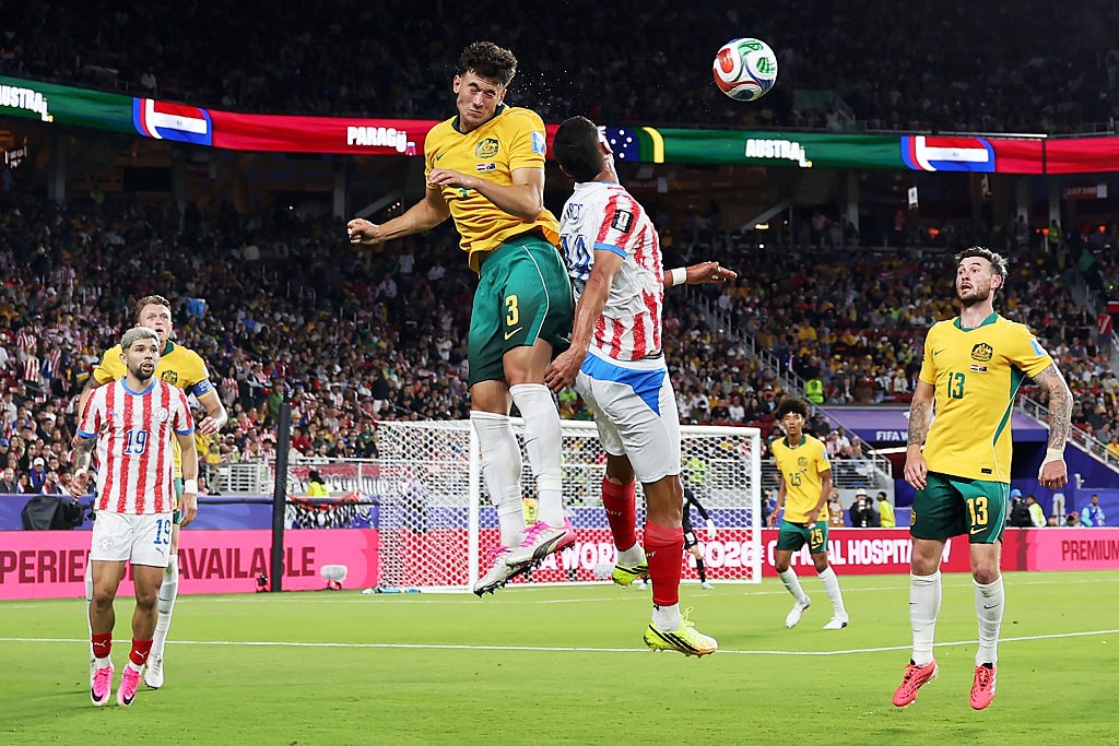 Dois jogadores de futebol saltam para cabecear a bola no centro de um campo verde, com um estádio lotado ao fundo. O jogador à esquerda veste camisa amarela e shorts verdes, o da direita, camisa listrada vermelha e branca com shorts azuis e brancos. A bola está no ar acima deles. Outros jogadores e torcedores são visíveis no fundo