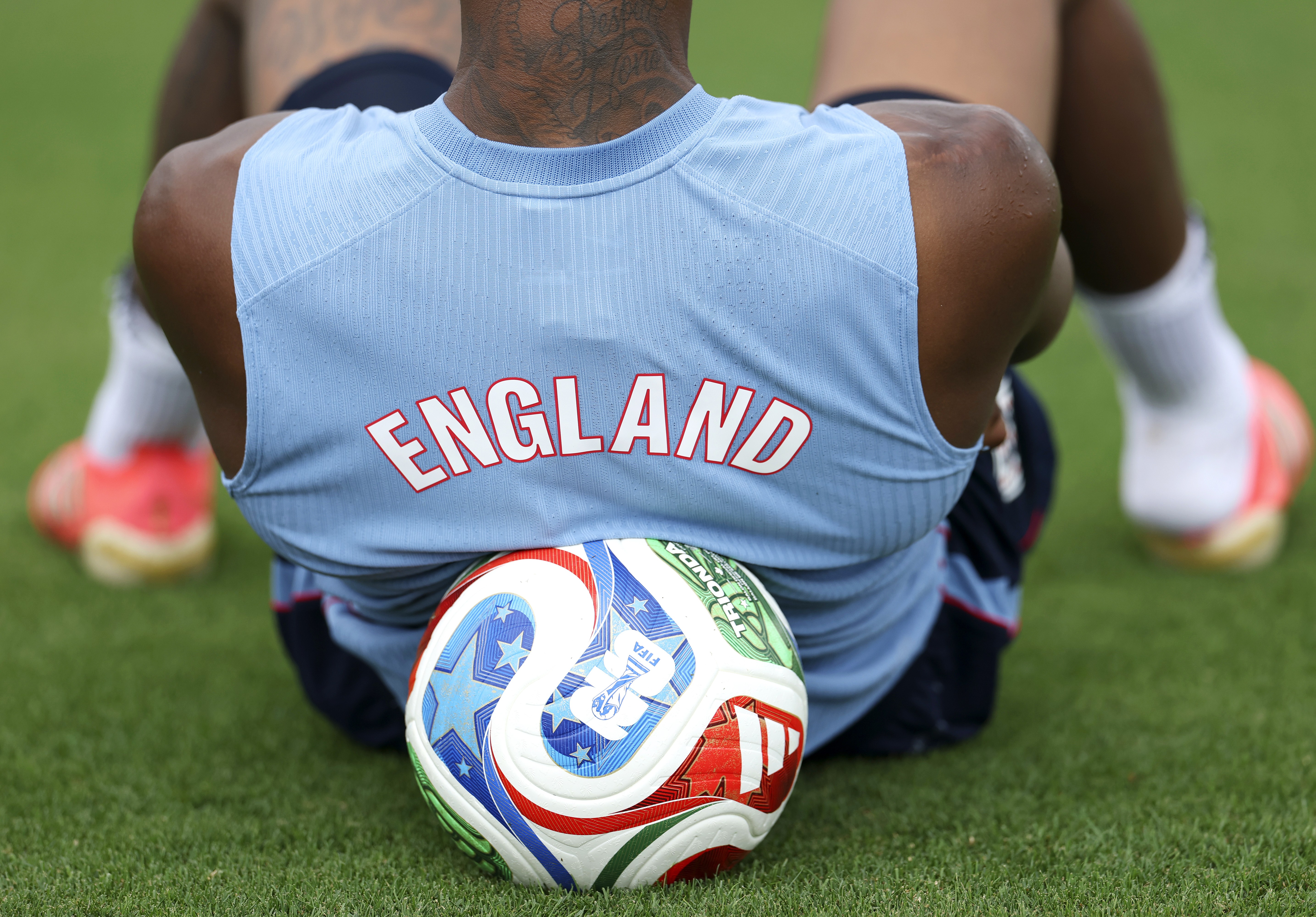 Jogador de futebol de costas, vestindo camisa azul clara com ENGLAND em vermelho, sentado no gramado com uma bola de futebol colorida entre as pernas