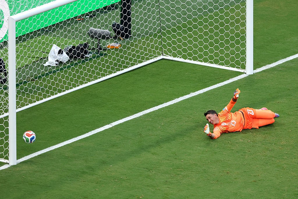 Goleiro de uniforme laranja deitado no gramado verde, com a bola colorida dentro do gol à esquerda, após uma defesa
