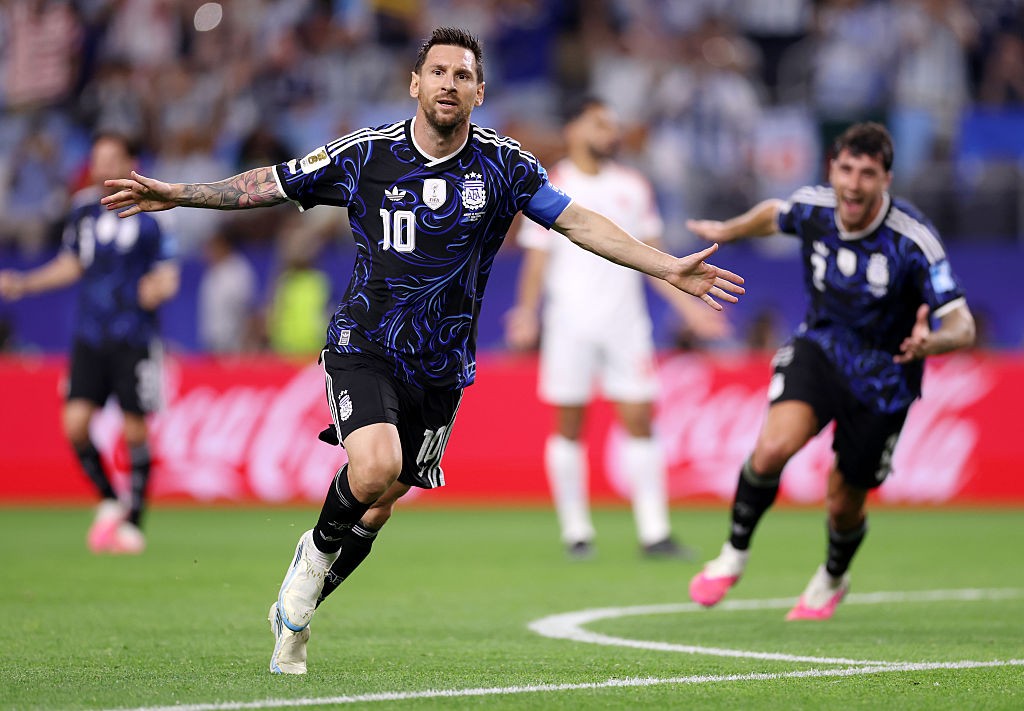 Lionel Messi, com barba e camisa 10 azul-marinho e preta, corre no campo de futebol com os braços abertos, celebrando um gol. Outros jogadores e torcedores desfocados ao fundo.