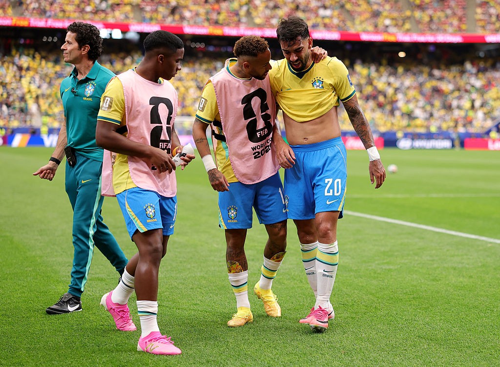 Três jogadores de futebol brasileiros, dois com coletes rosa sobre a camisa amarela e um sem camisa, caminham no campo verde. Um quarto homem, de uniforme verde, os acompanha.
