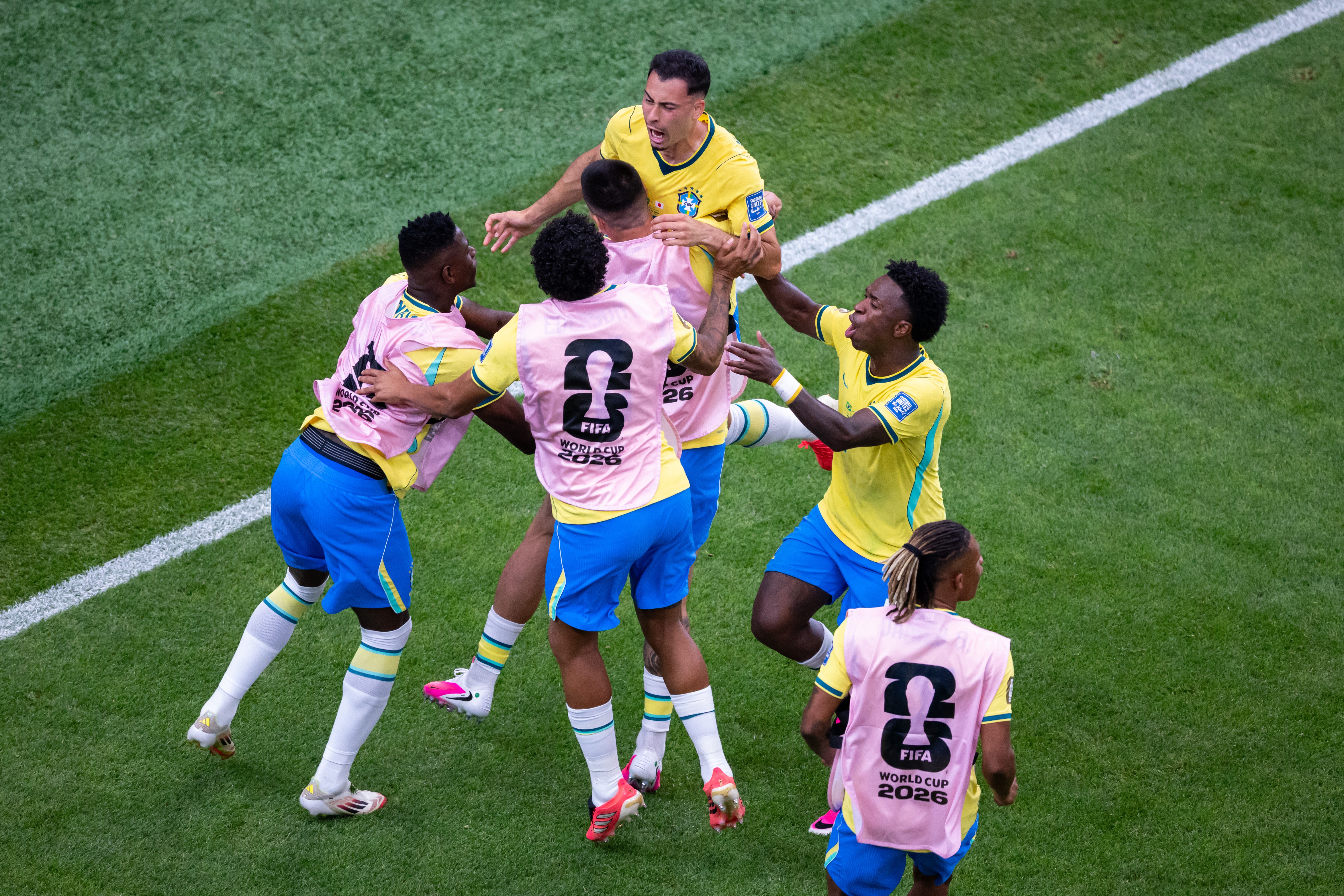 Jogadores de futebol brasileiros, vestindo camisas amarelas e coletes de treino rosa, se abraçam e comemoram em campo de grama verde, com uma linha branca visível