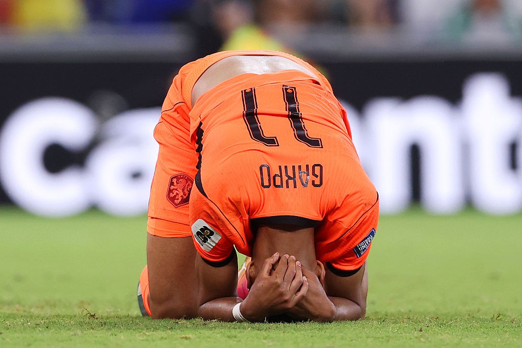 Jogador de futebol com camisa laranja e número 11, nome GAKPO, ajoelhado no gramado com a cabeça baixa e as mãos no rosto, expressando desespero ou frustração. Ao fundo, letras brancas desfocadas de um letreiro publicitário