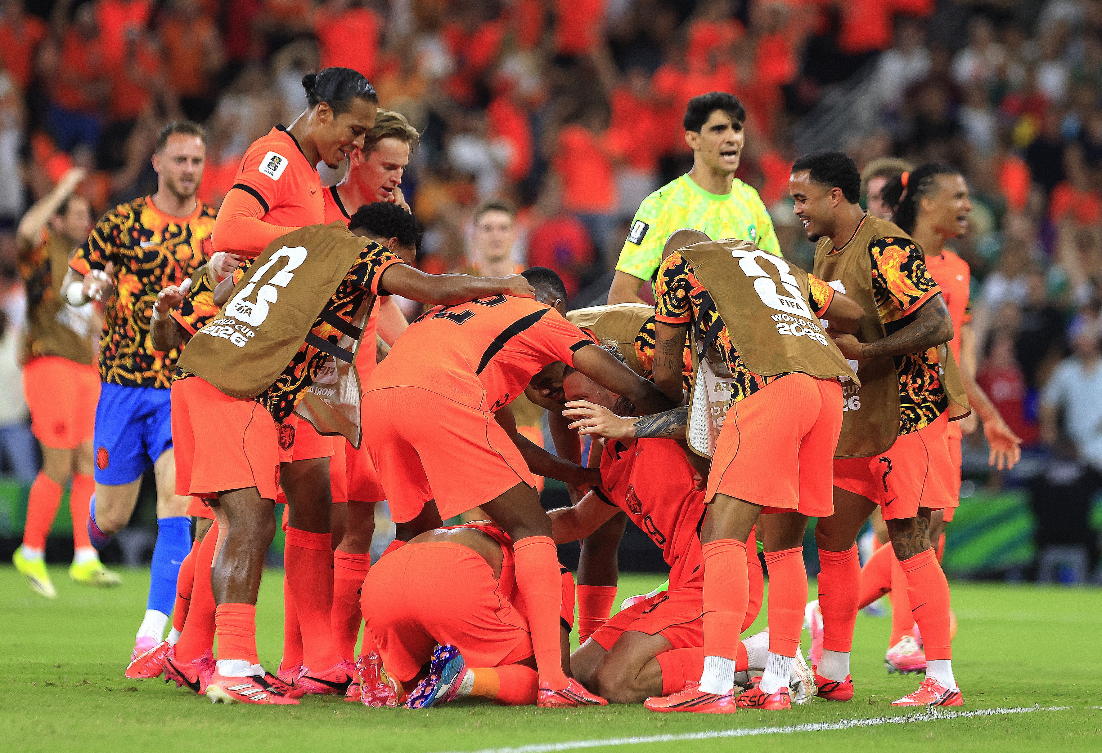 Jogadores de futebol com uniformes laranja e pretos, alguns com coletes marrons, se abraçam em comemoração no campo verde de um estádio lotado