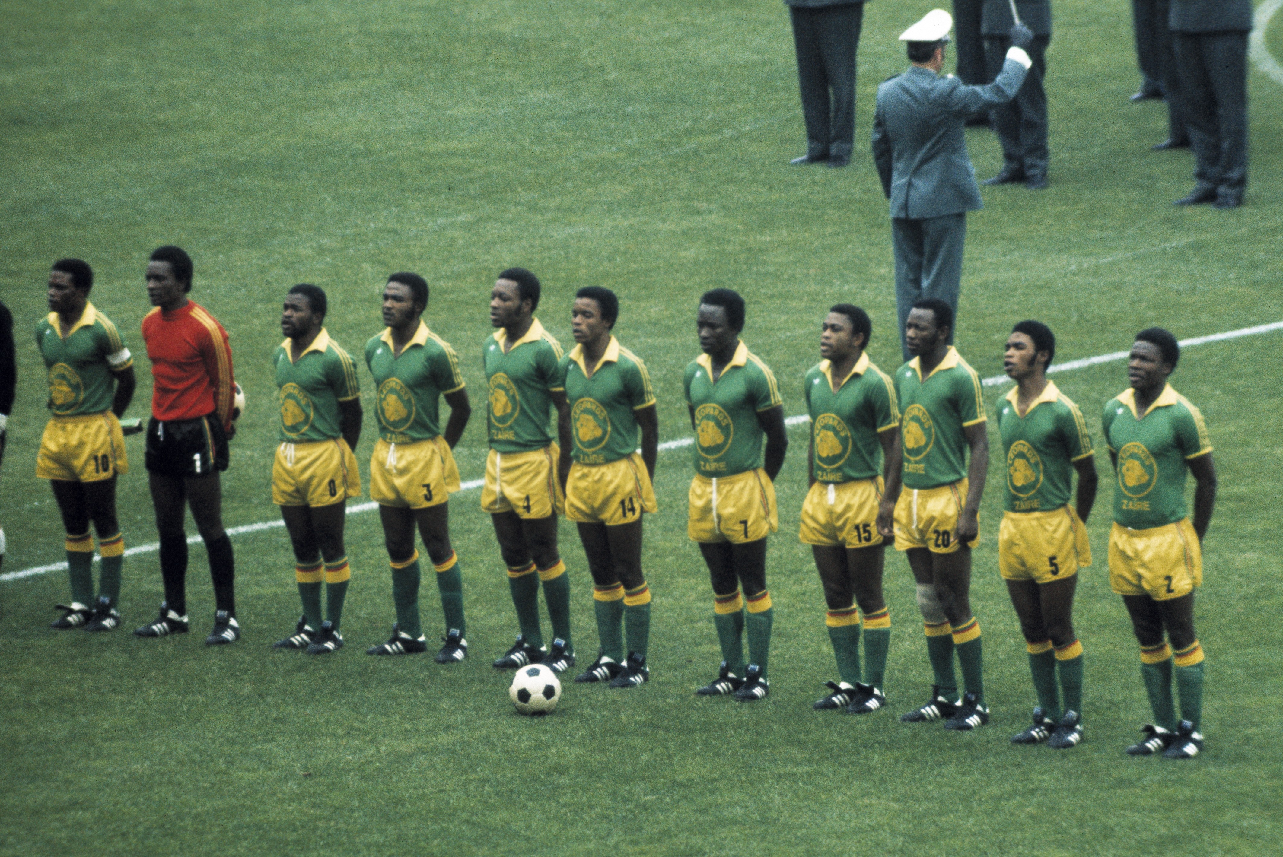Time de futebol masculino do Zaire, com 11 jogadores negros em campo, vestindo camisas verdes com detalhes amarelos e shorts amarelos, alinhados em pé sobre a grama verde de um estádio, com uma bola de futebol no chão. Ao fundo, oficiais de terno e um homem de uniforme militar branco e azul.